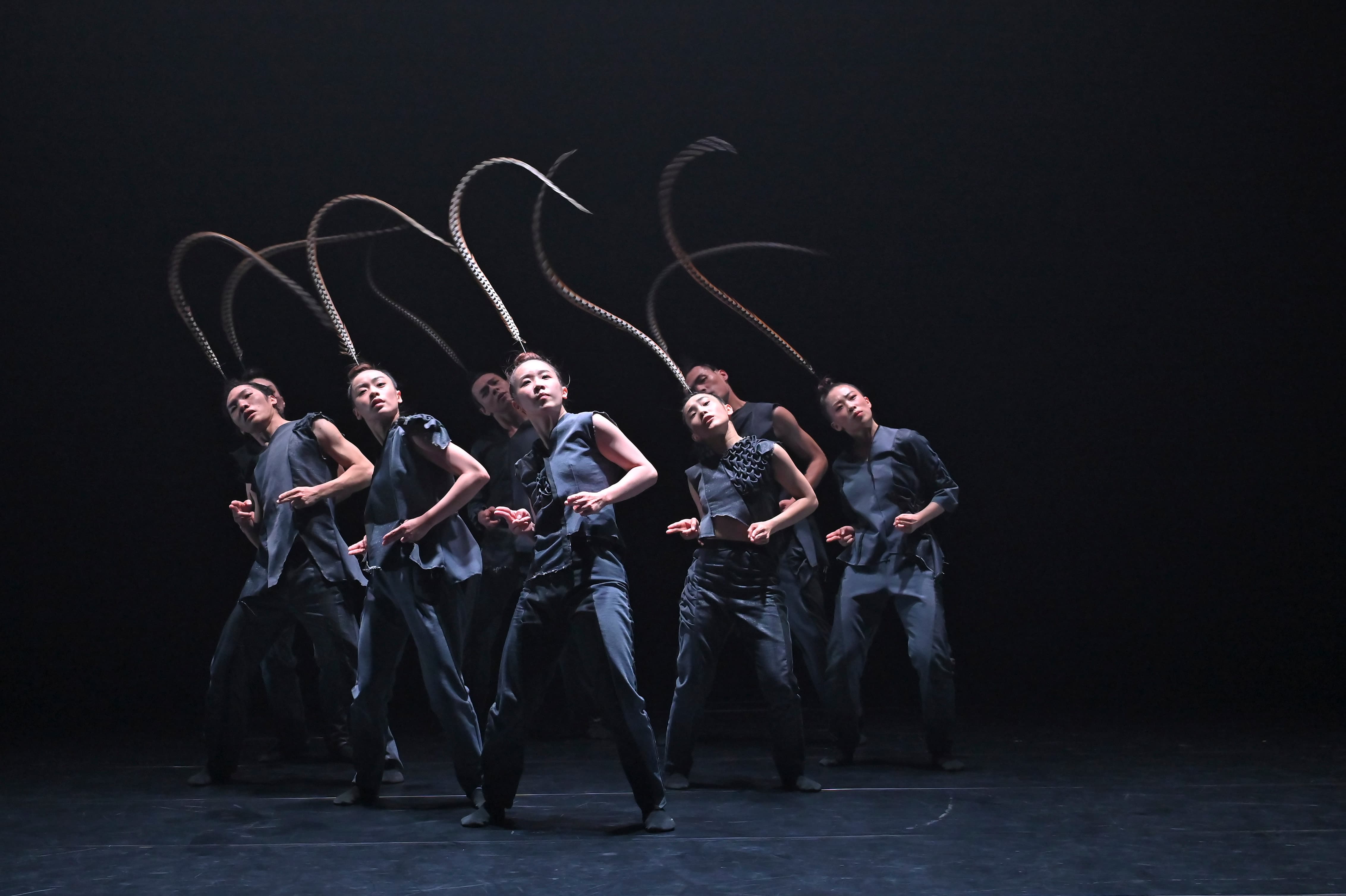 a group of 7 asian dancers on stage, wearing black clothes adn a long bird feather on their heads 