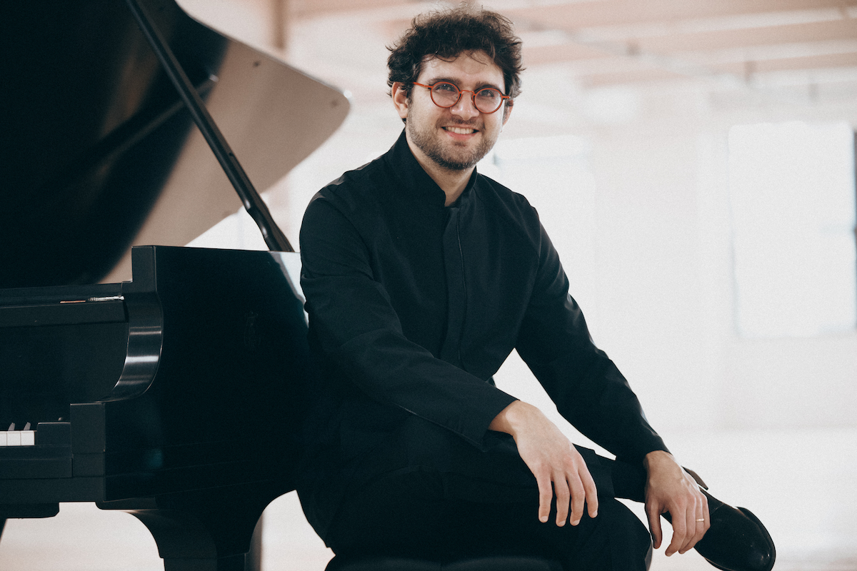 Roman Robinavich is wearing a black shirt, black pants, and glasses. He is seated in front of a grand piano.