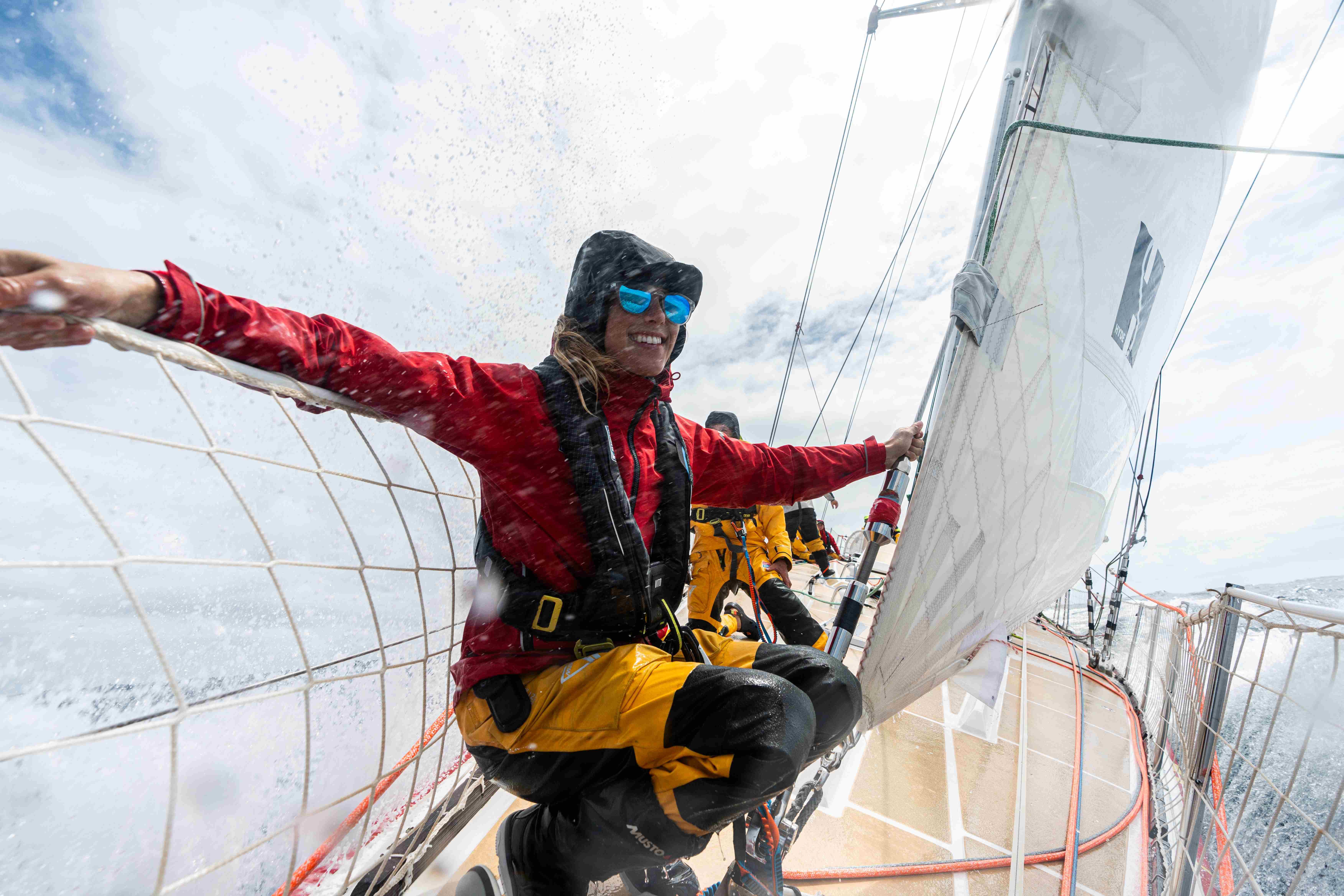 A young woman wearing storm-worthy sailing kit smiles as she crouches and holds onto the deck rail of an ocean sailing vessel. Ocean spray hits her from behind and other crew can be seen behind her.