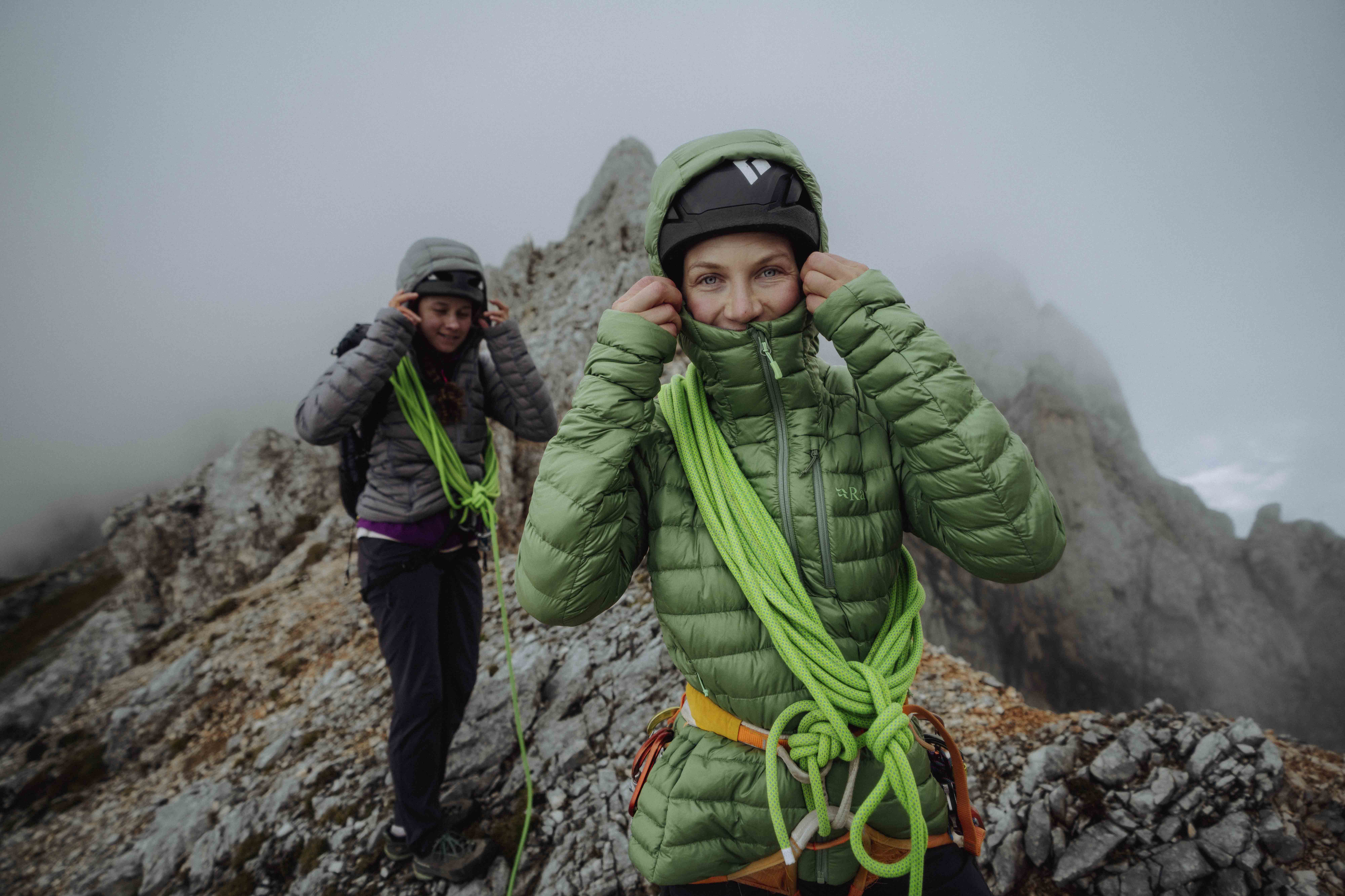 Two women, walking on a mountain ridge inside a cloud, pull up the hoods of their green Rab down jackets against the weather. They wear a rope mountaineer coiled to both and are smiling.