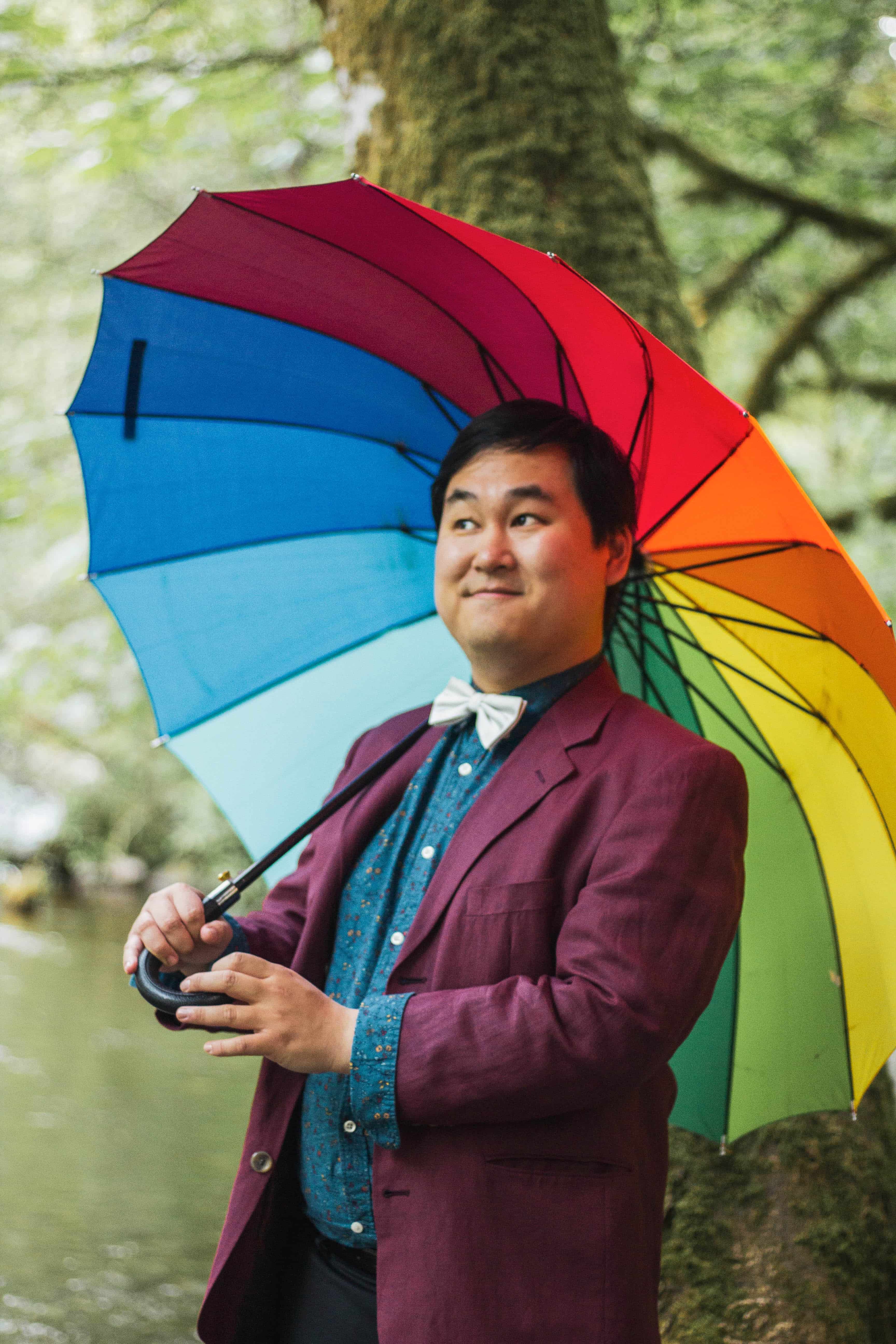Man holding rainbow coloured umbrella open behind him and smiling offside to the camera