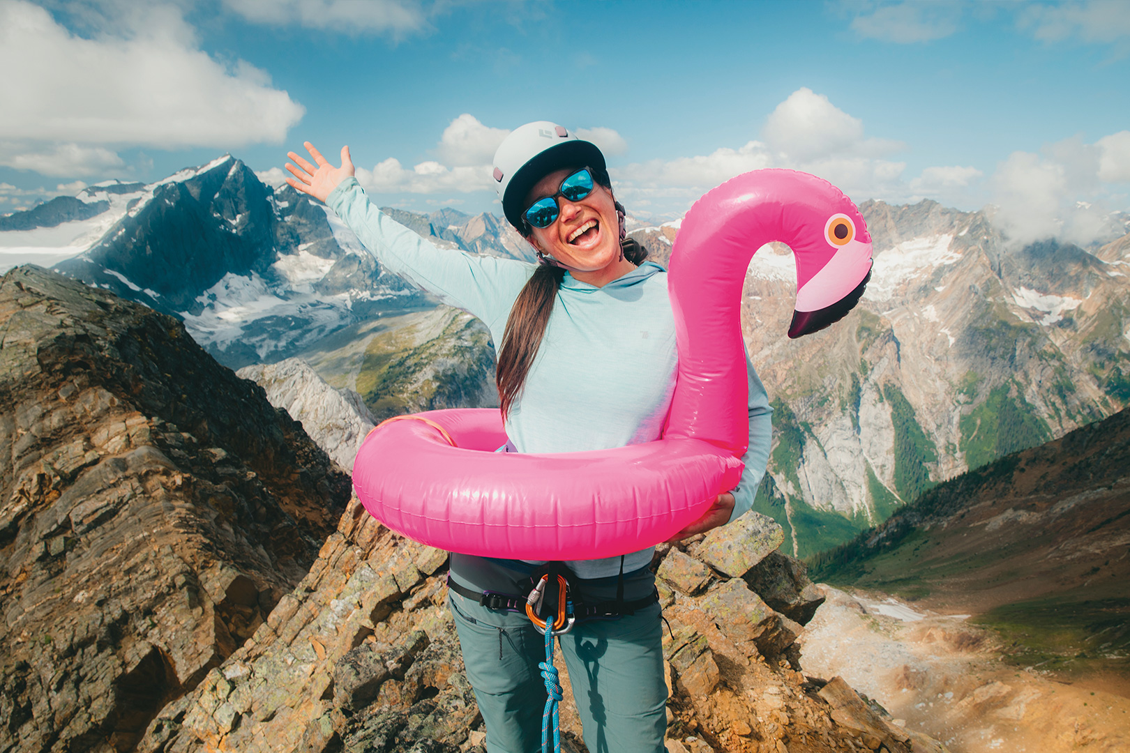 A woman in climbing gear stands on a summit. One handis raised in the air and the other holds up a pink flamingo inflatable ring around her waist. 