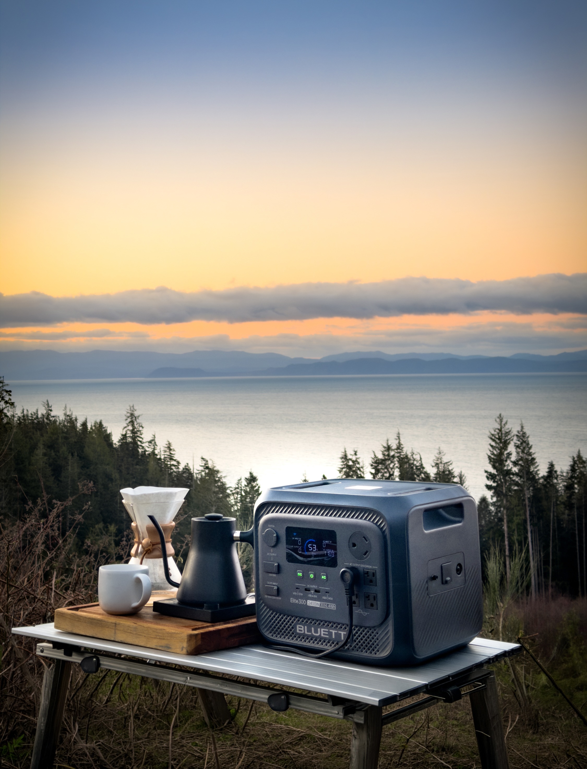 A small generator connected to a kettle and pour over coffee sit on a camp table in front of a mountainous coastal sunrise scene.