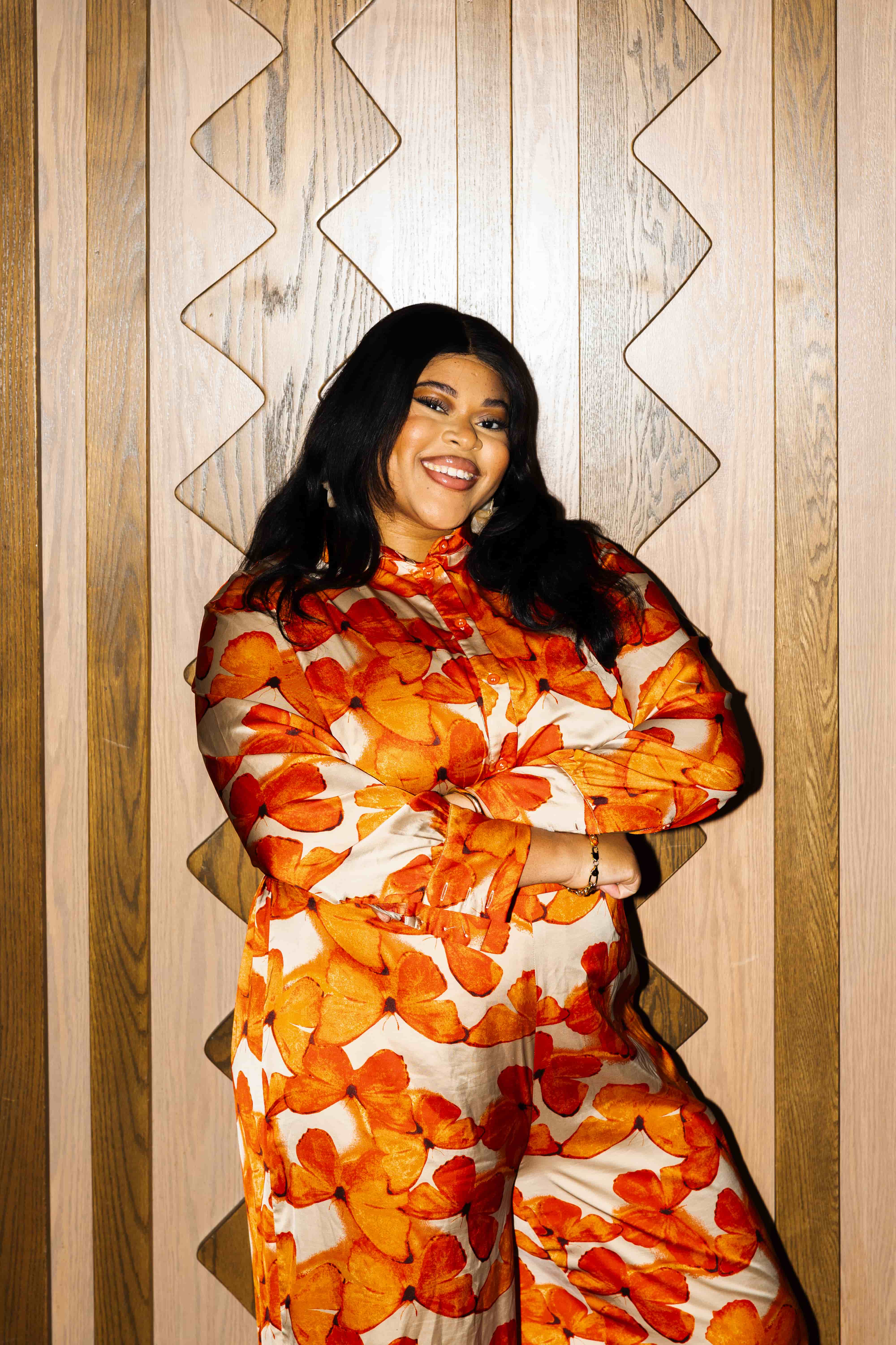 A smiling person with long dark hair poses confidently with crossed arms, wearing a flowing outfit covered in bold orange floral patterns. They stand against a light wood-paneled wall with geometric detailing, creating a warm and vibrant portrait