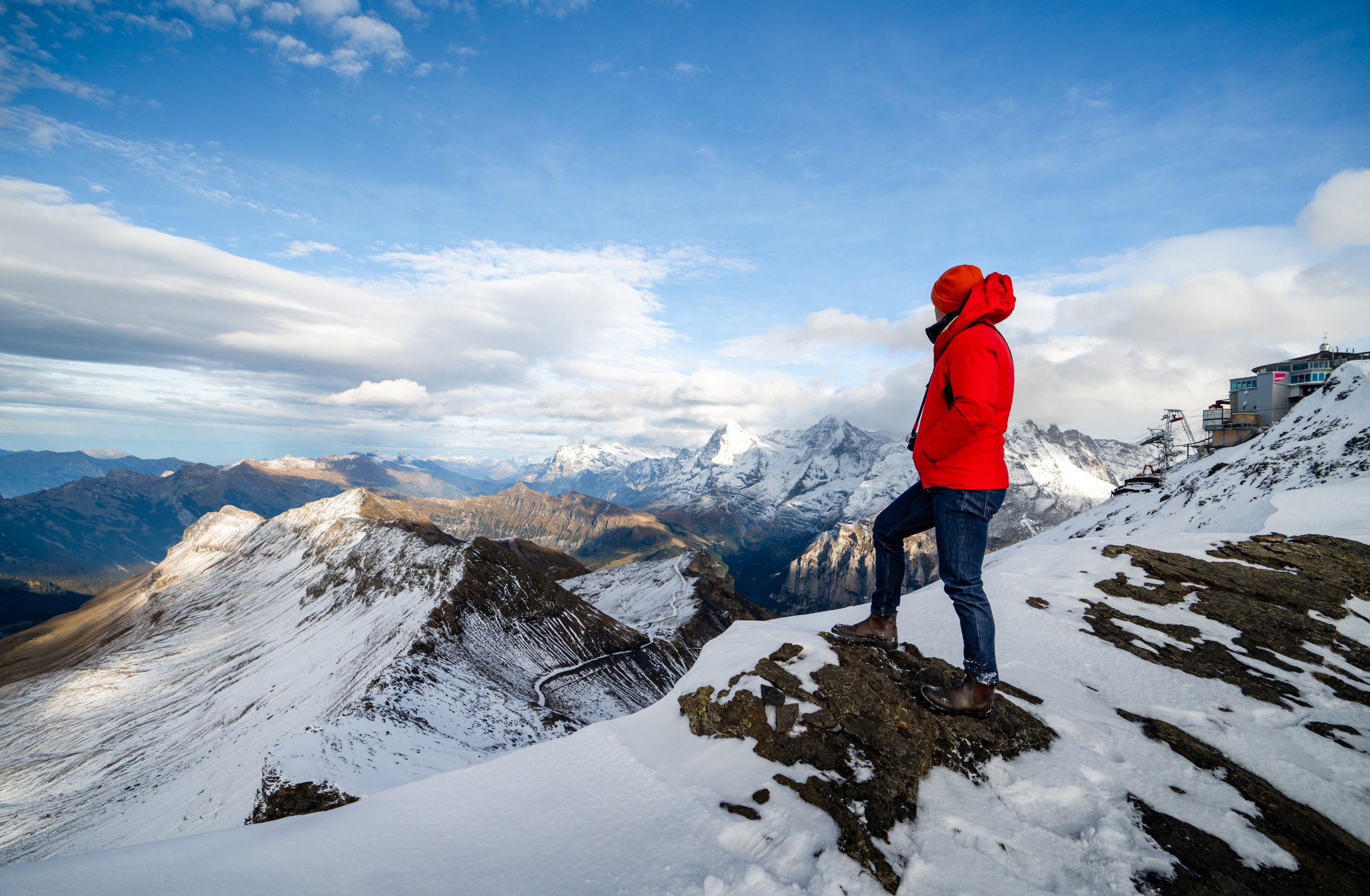 A man in a red jacket, red hat, and denim pants stands lookin out over lightly snow peaks of the Swiss Alps