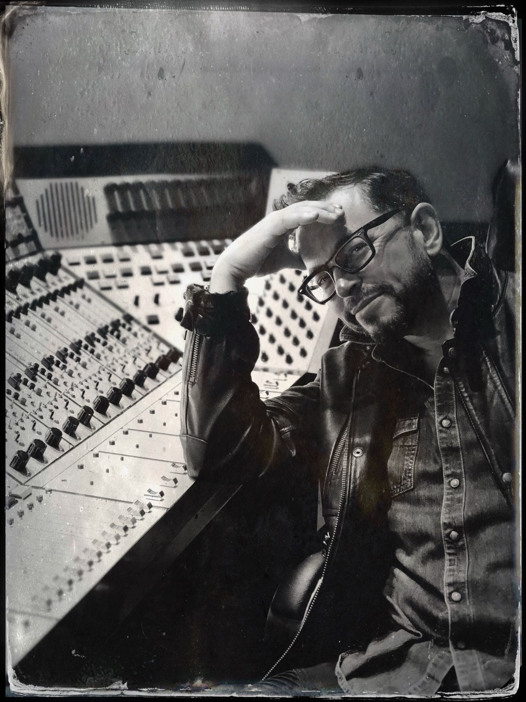black and white portrait of a man with glasses seating next to a mixing console.