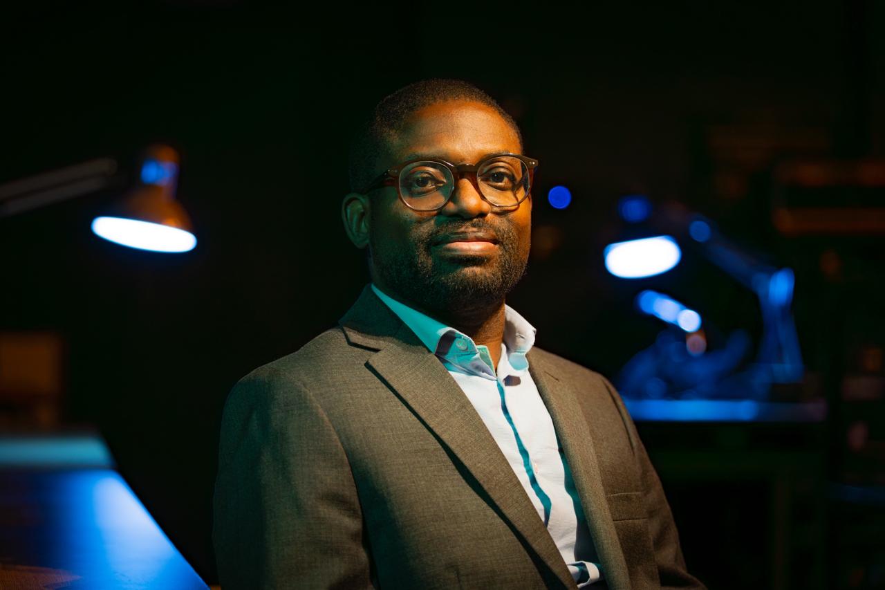 Dr Taiwo Afolabi, wearing a suit jacket and glasses, gazes directly into the camera