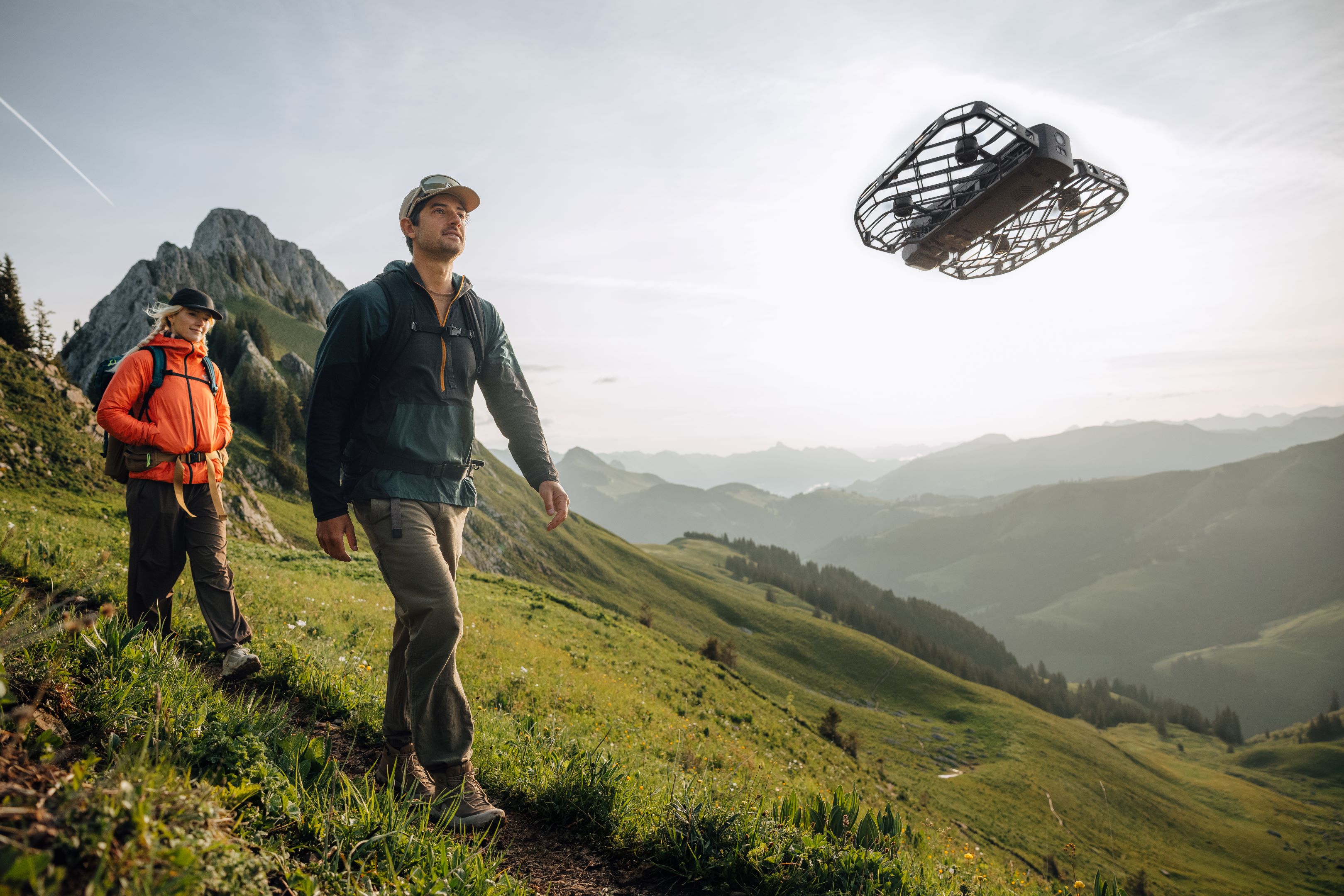 A woman and a man walk down a narrow dirt trail on the grassy slope of a mountainside with a drone flying in front of them.