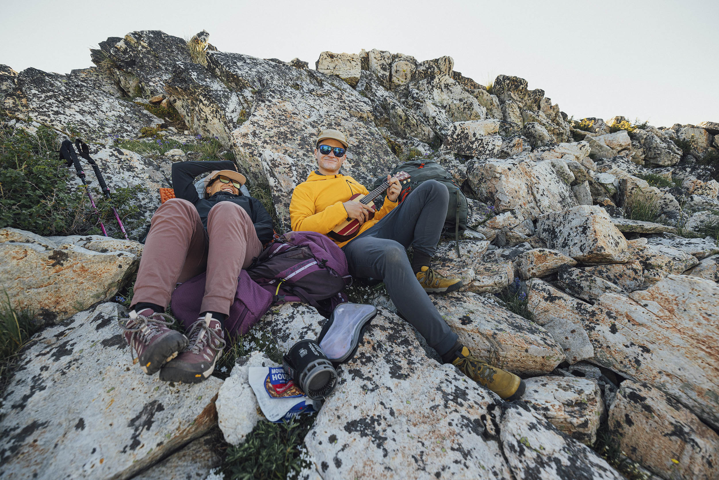 Two folks sit on the side of a rocky trail in Fall seasonal backpacking clothing. They have a camp stove in front of them, and one holds a ukulele and is smiling at the camera.