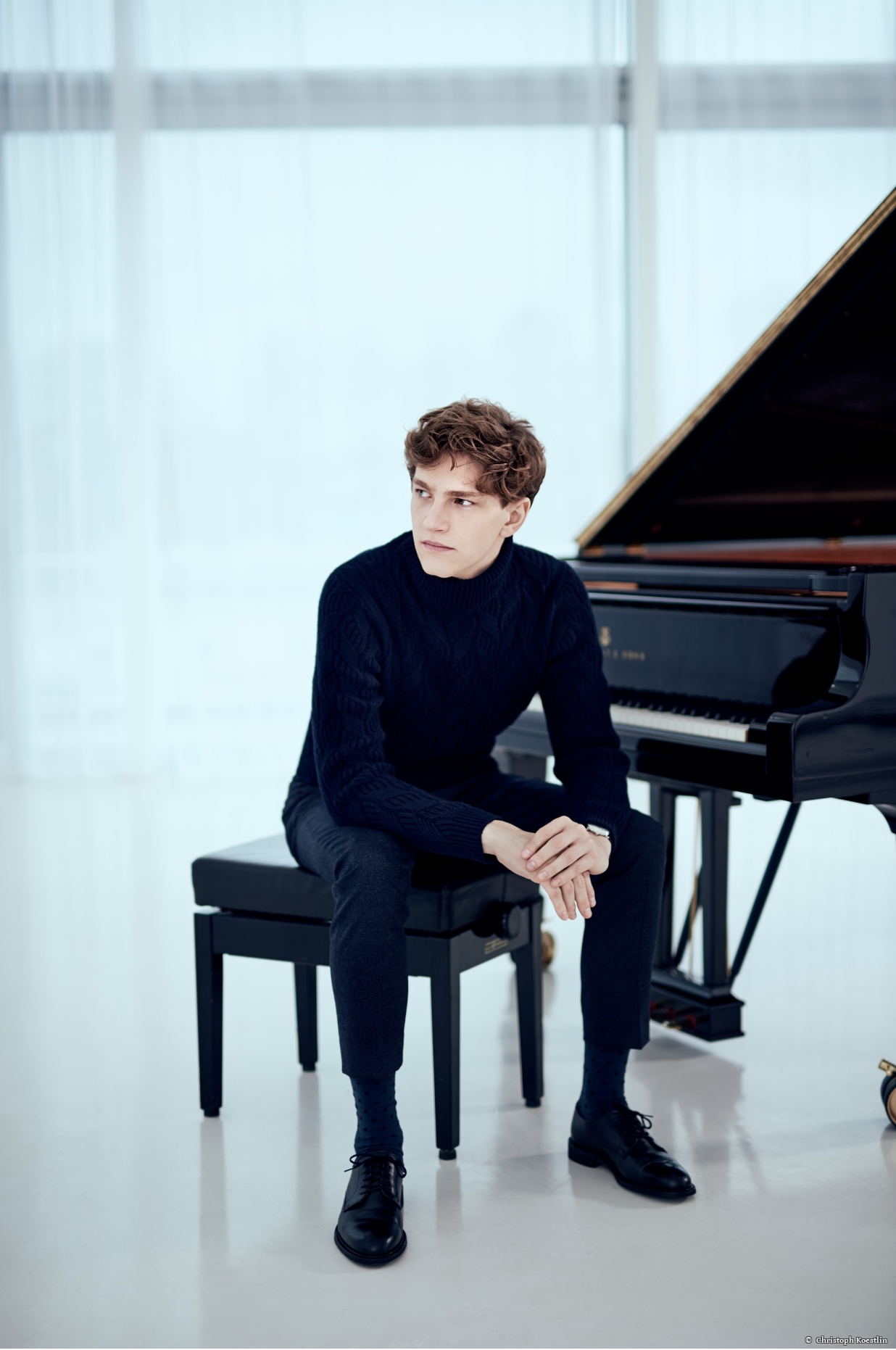 Jan Lisiecki is seated on a piano bench next to a grand piano