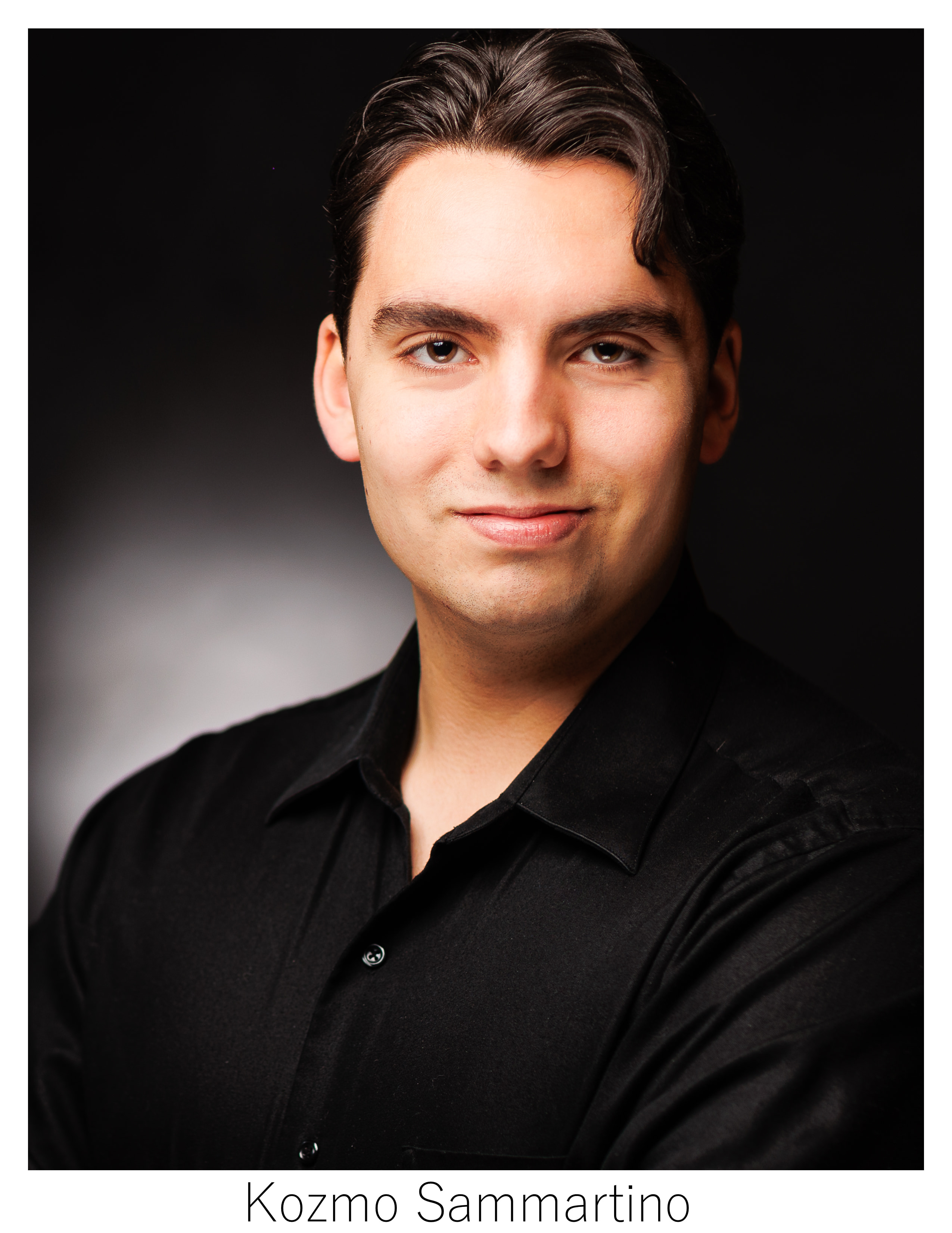 Man in black standing against dark background smiling lightly at camera with short wavy brown hair