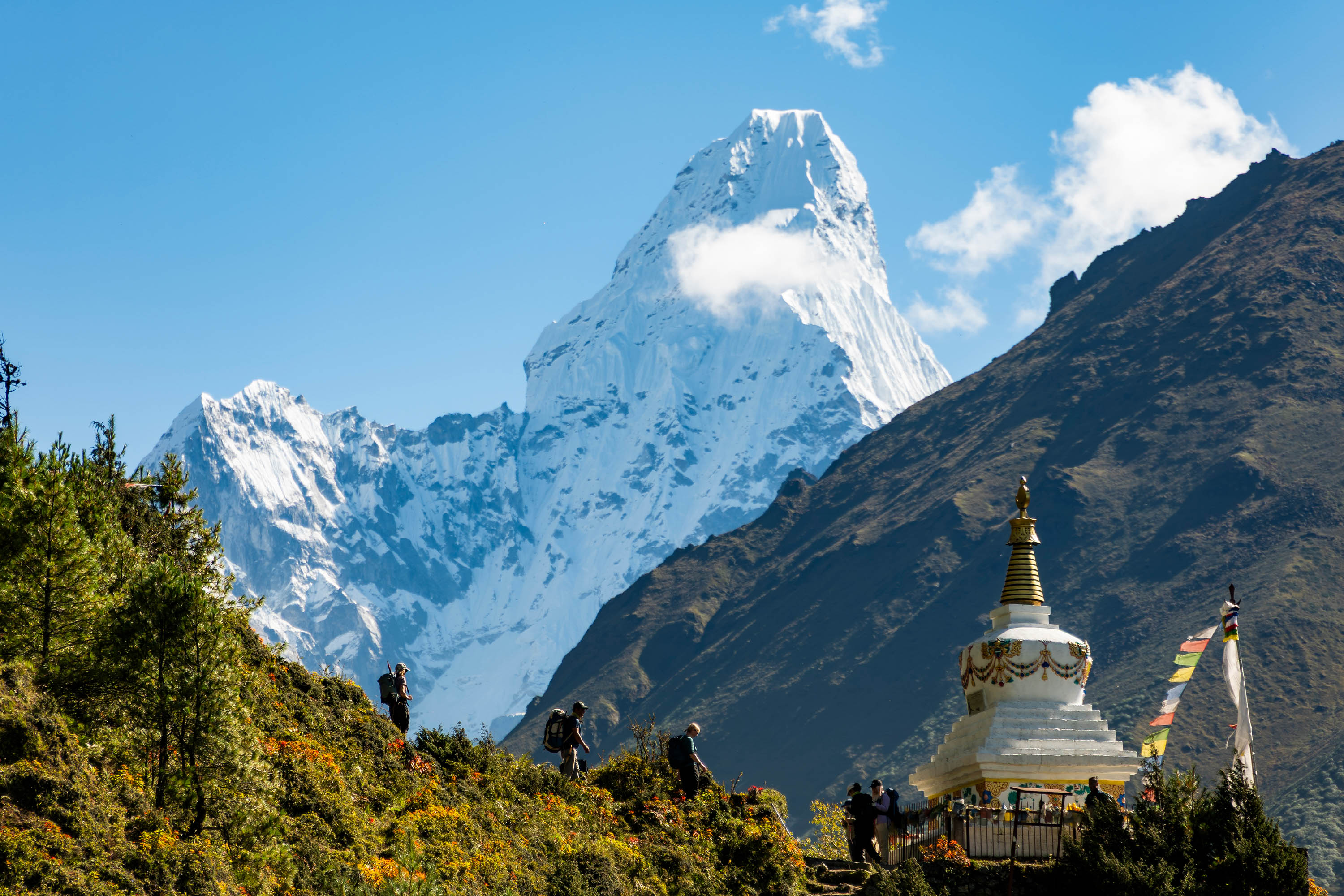The pointed peak of Ama Dablam juts skyward behind two ridges. In the foreground a group of people hike by an ornate Buddhist stupa religious site.