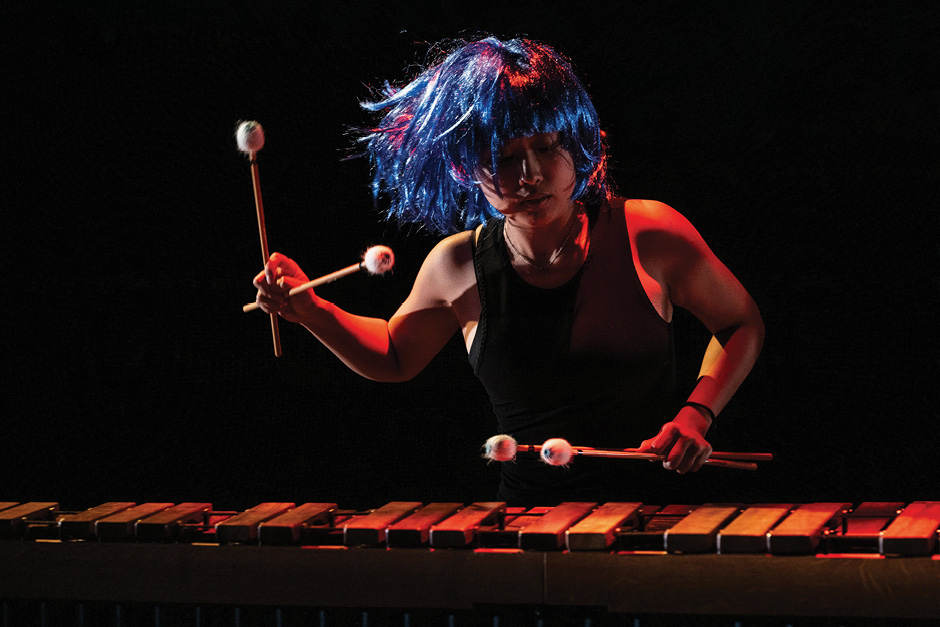 A musician in a blue wig plays a percussion instrument