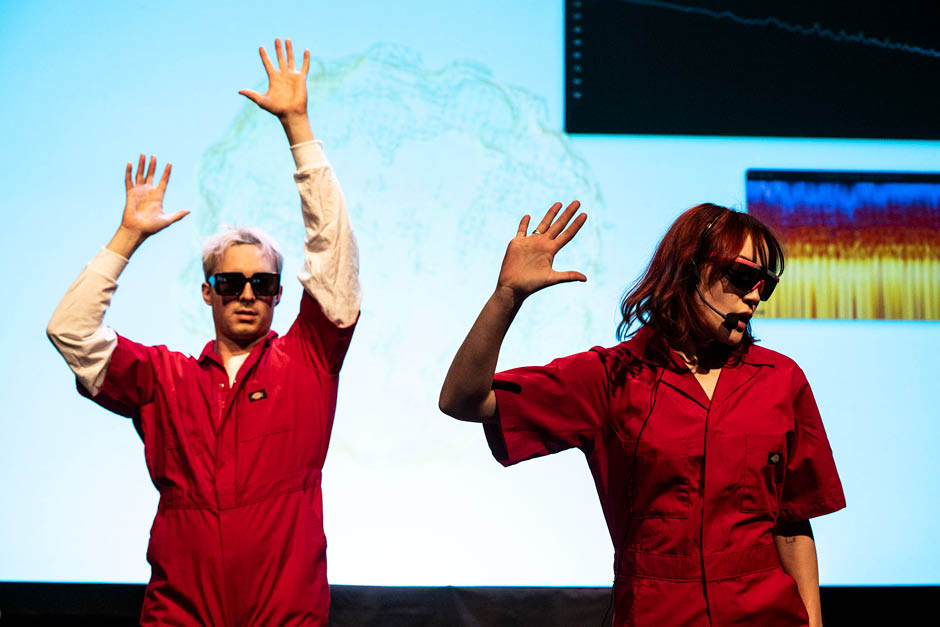 Two musicians in bright red coveralls perform in front of a blue backdrop