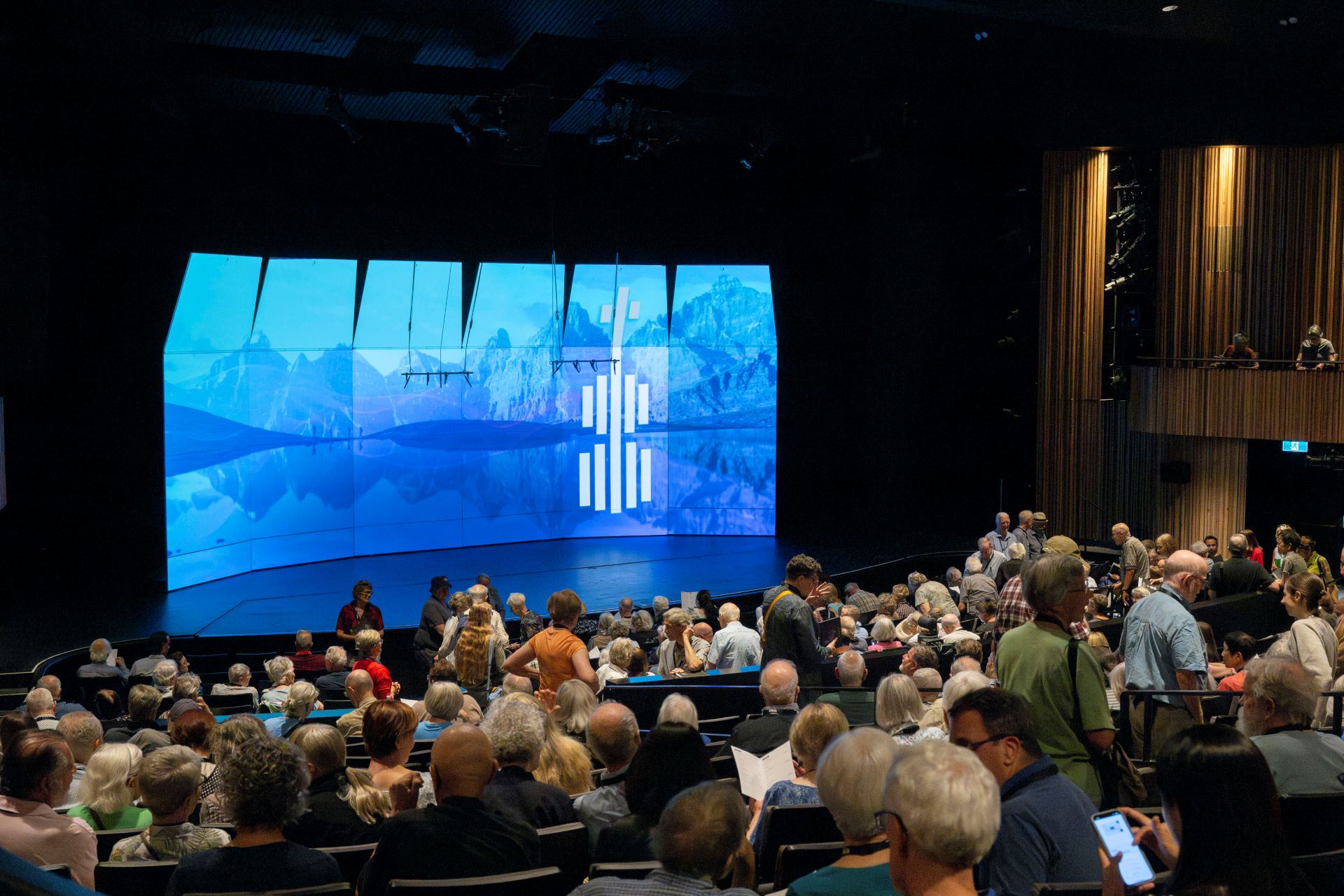 Interior Jenny Belzberg Theatre - Packed Audience in front of empty stage and blue screen with BISQC logo