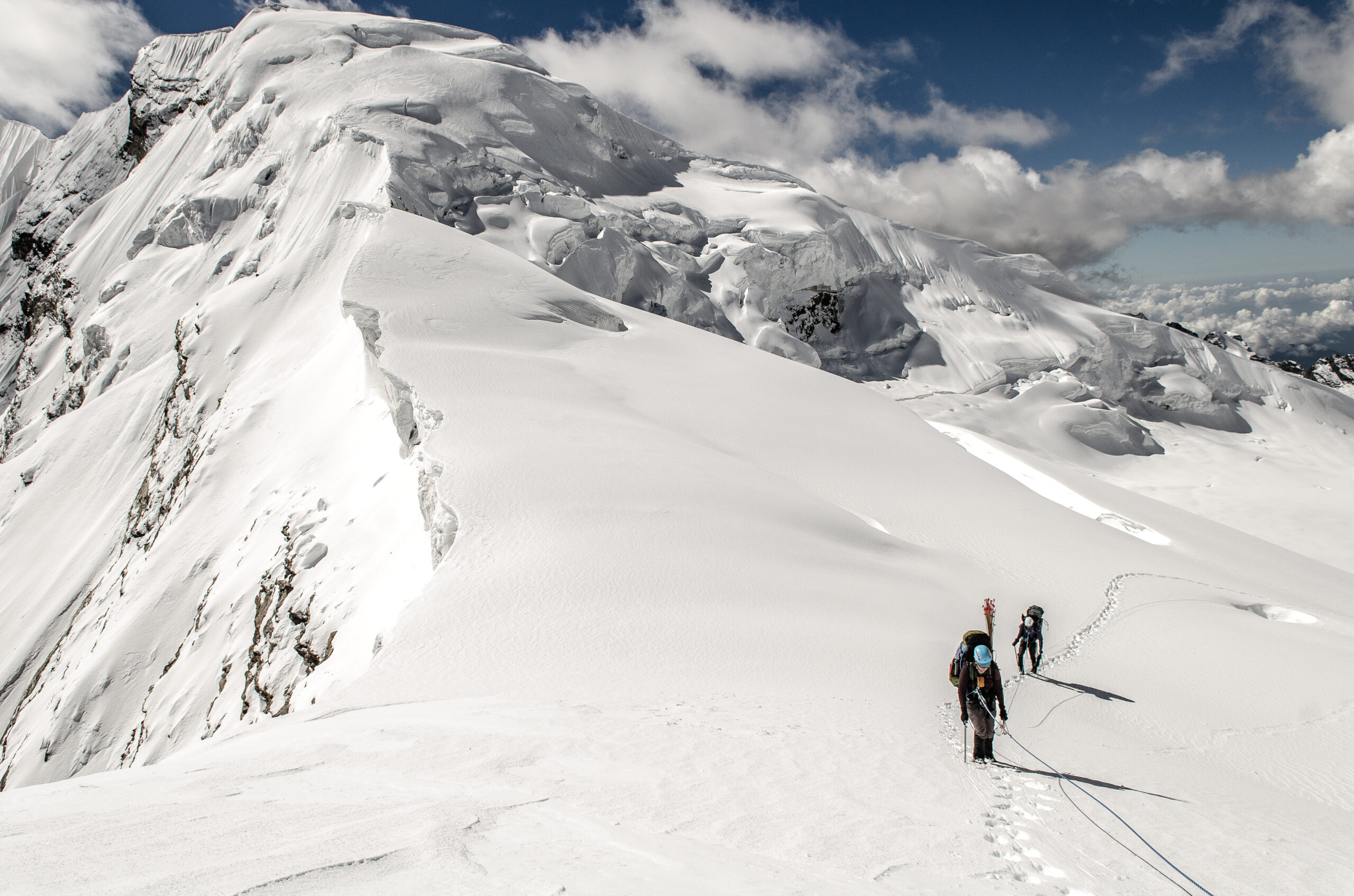 A group of mountaineers connected by a rope walk a snow ridge with a heavily glaciate peak close behind them.