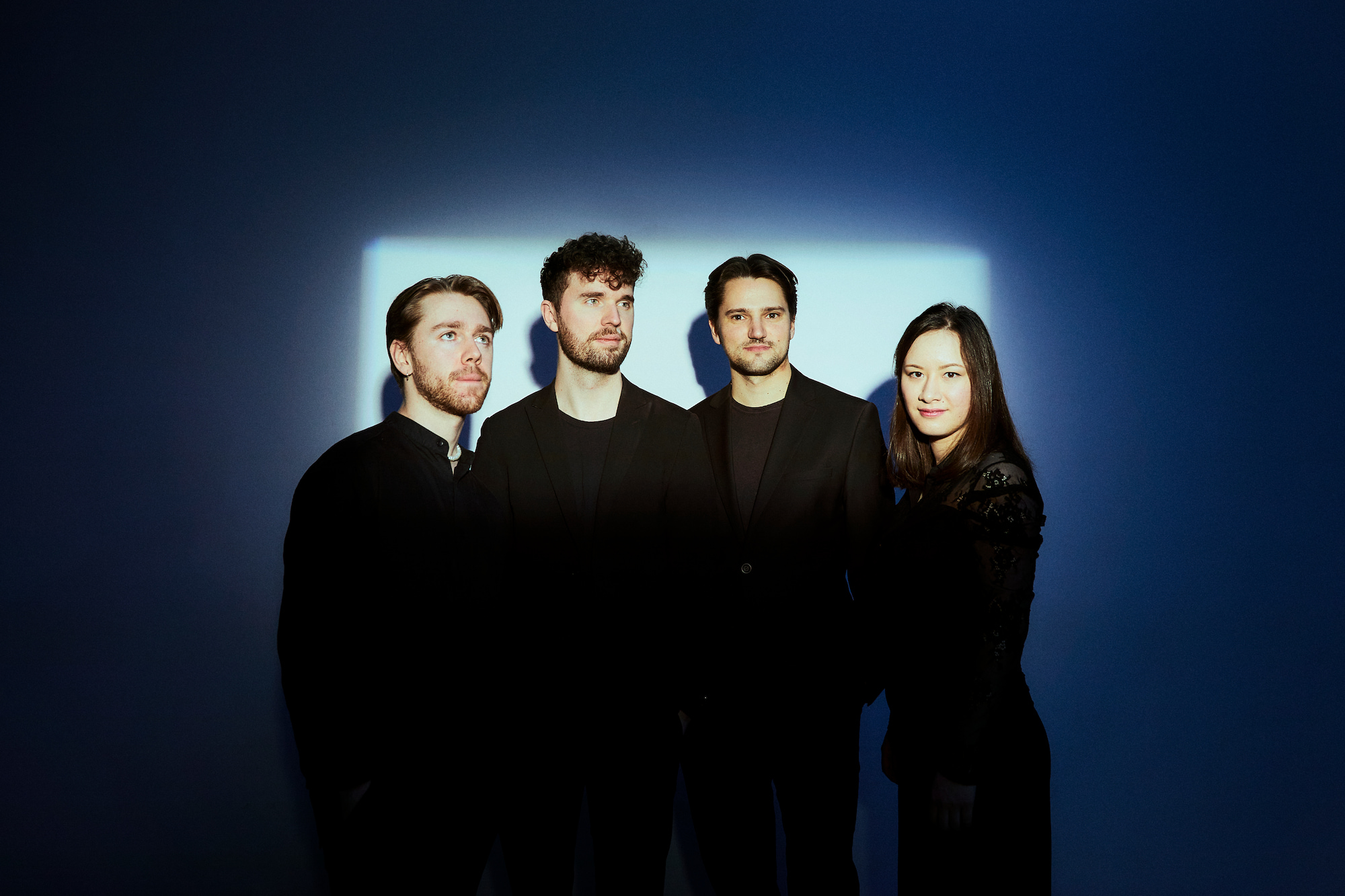 Four people dressed in black are standing in front of a blue wall