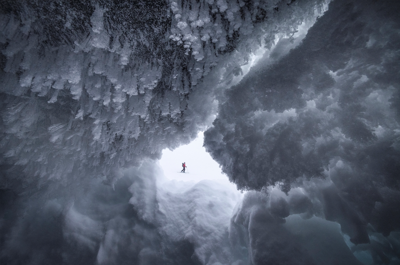 A person in glacier skiing equipment crosses in front of  a frost covered cave from which the photo is taken inside of.