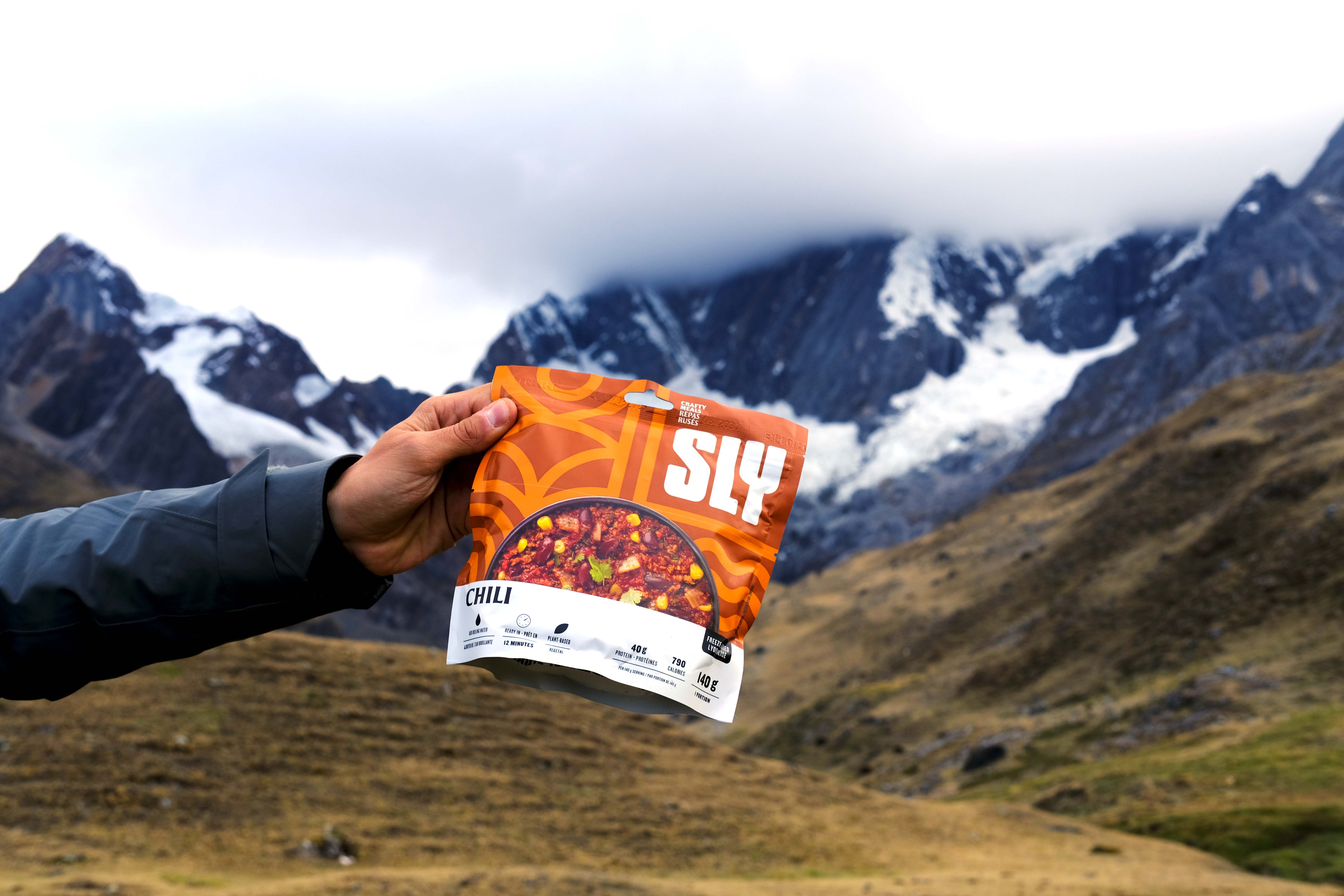 From the left sid eof the image frame a hand holds out and orange dehydrated meal bag of SLY Foods in front of an out of focus glaciated mountain.