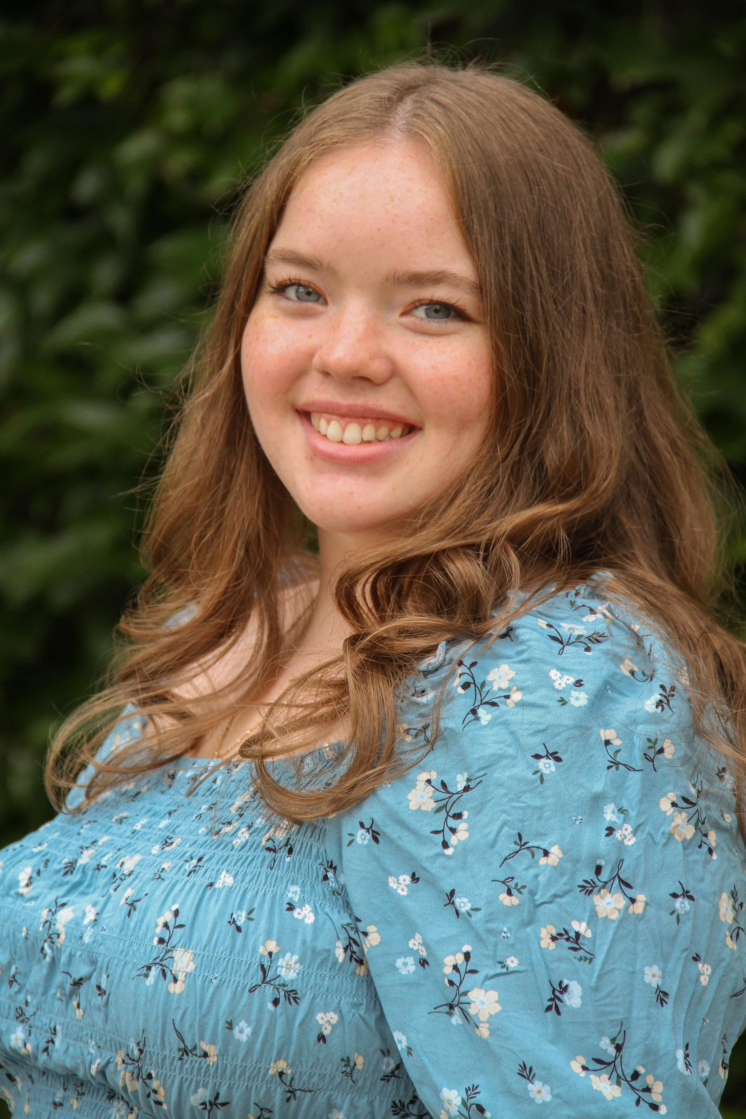 Woman with blue floral dress smiling directly at camera with light brown hair falling over shoulders