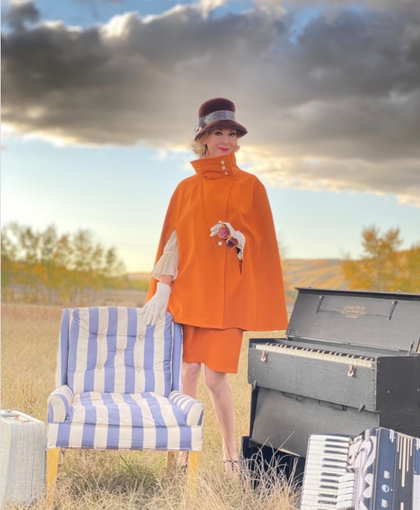 A person dressed in a vintage-inspired orange cape outfit and hat stands outdoors in a grassy field beside an upright piano, accordion, striped chair, and suitcase. Dramatic clouds and warm sunset light create a whimsical, cinematic atmospher