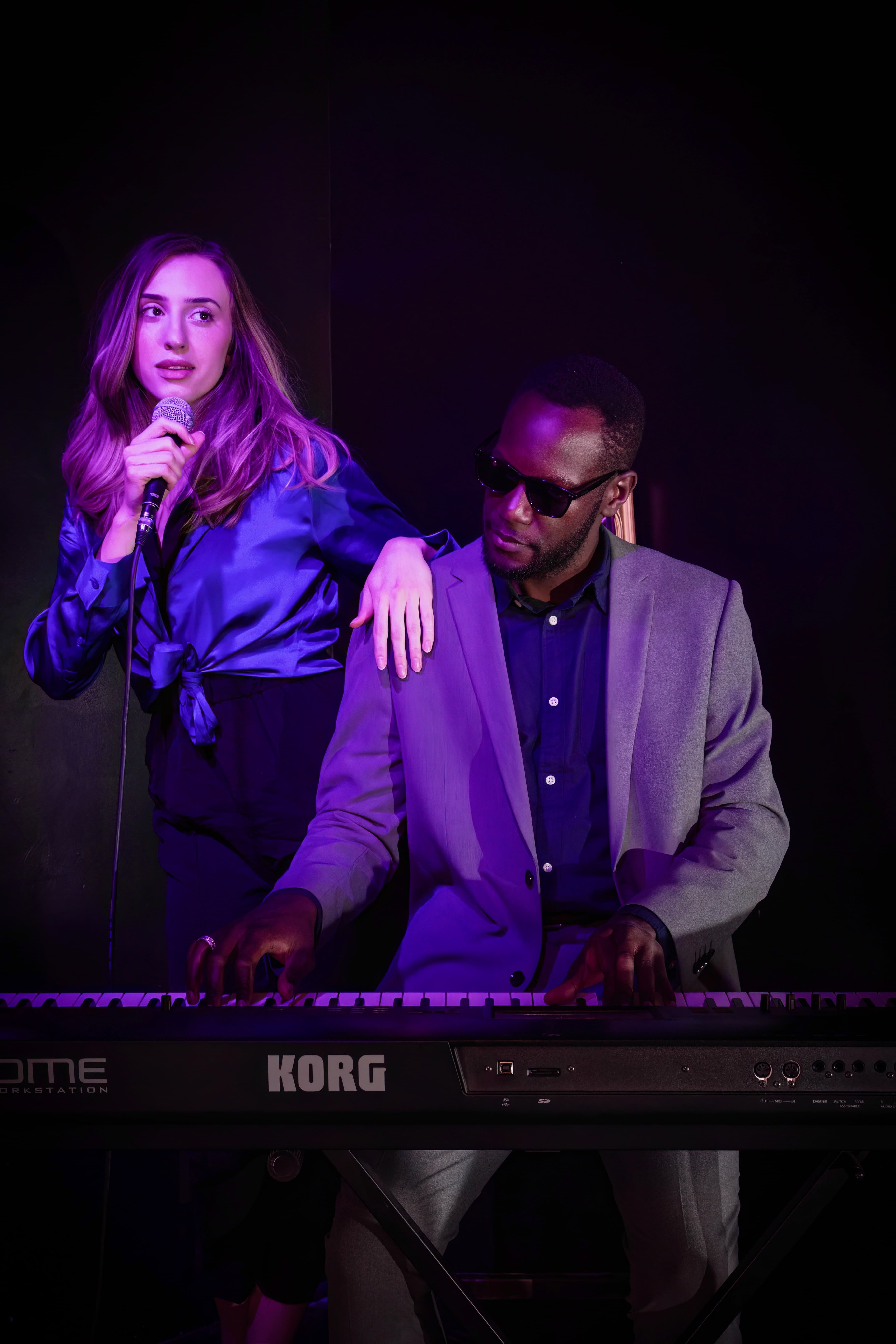 A singer holding a microphone stands beside a keyboard player performing under purple stage lighting. The keyboard player, wearing sunglasses and a light gray suit, plays a Korg keyboard while the singer leans casually on their shoulder in an intimate live music setting