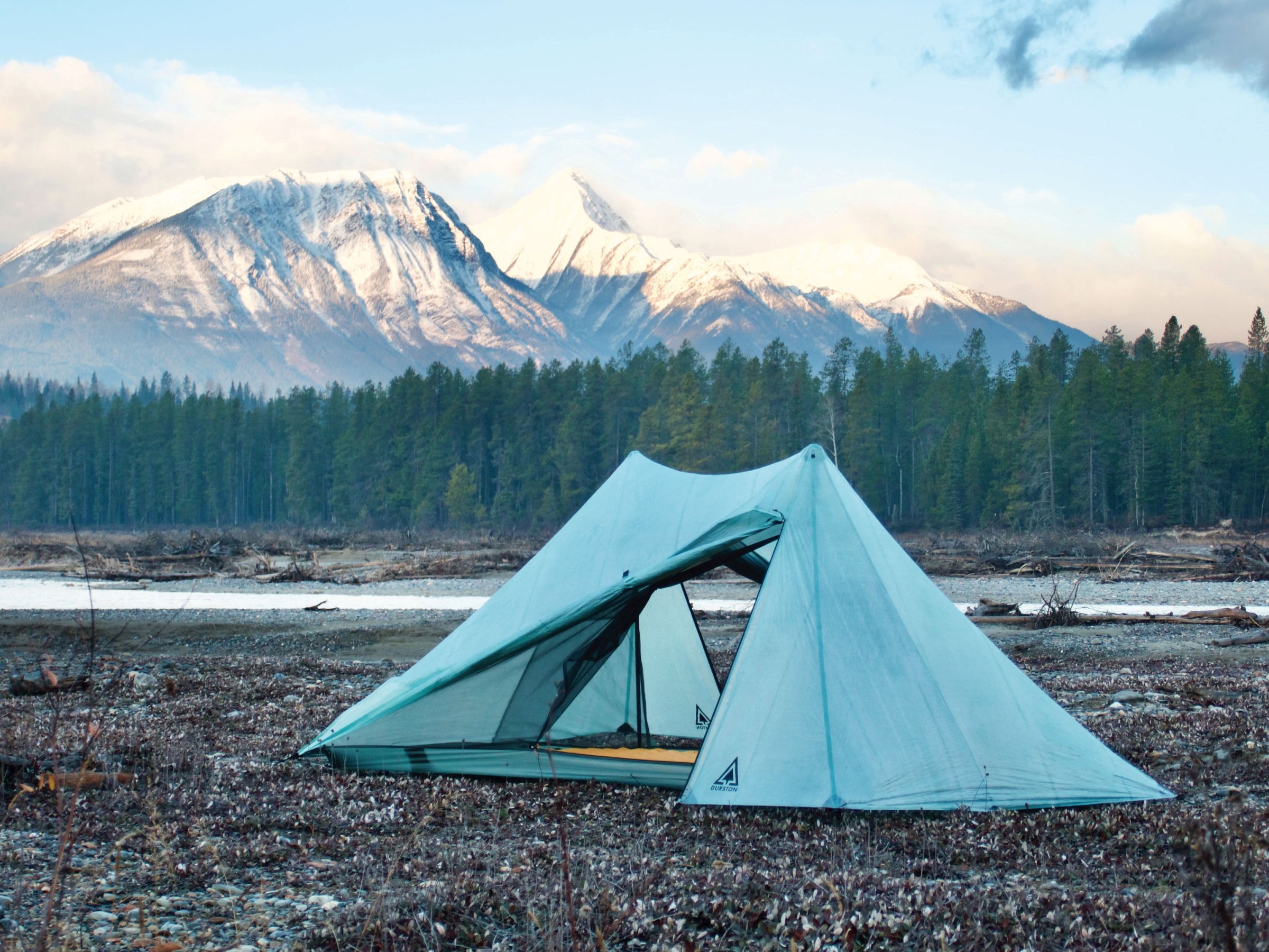 A Durston X-Mid Pro 1 tent is set up in gravel flats by a riverside. Snow covered late Fall peaks loom in the background.
