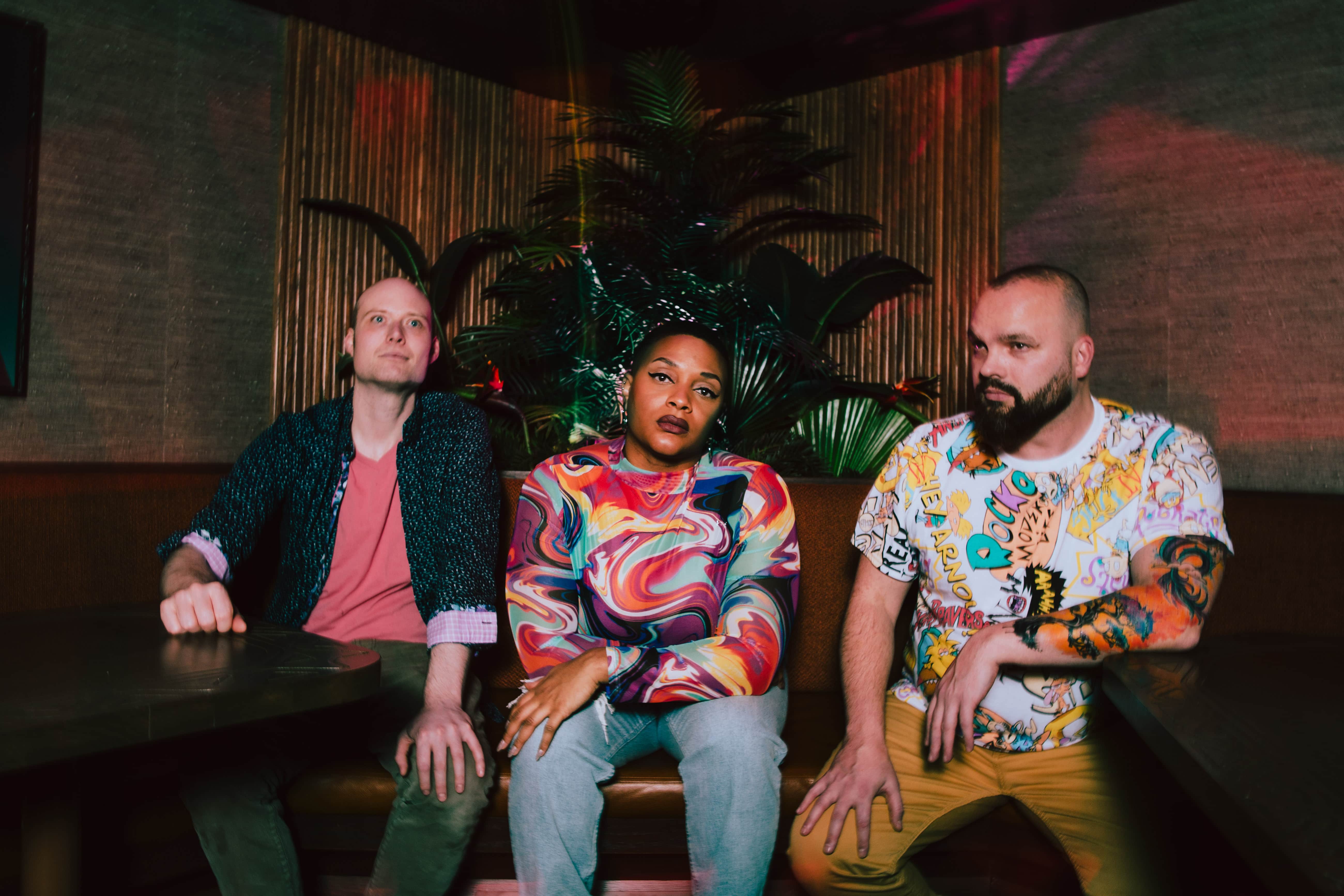 Three artists seated in a moody lounge setting with dramatic neon lighting and tropical plants in the background. The group wears colorful, expressive clothing and faces the camera with a bold, contemporary aesthetic