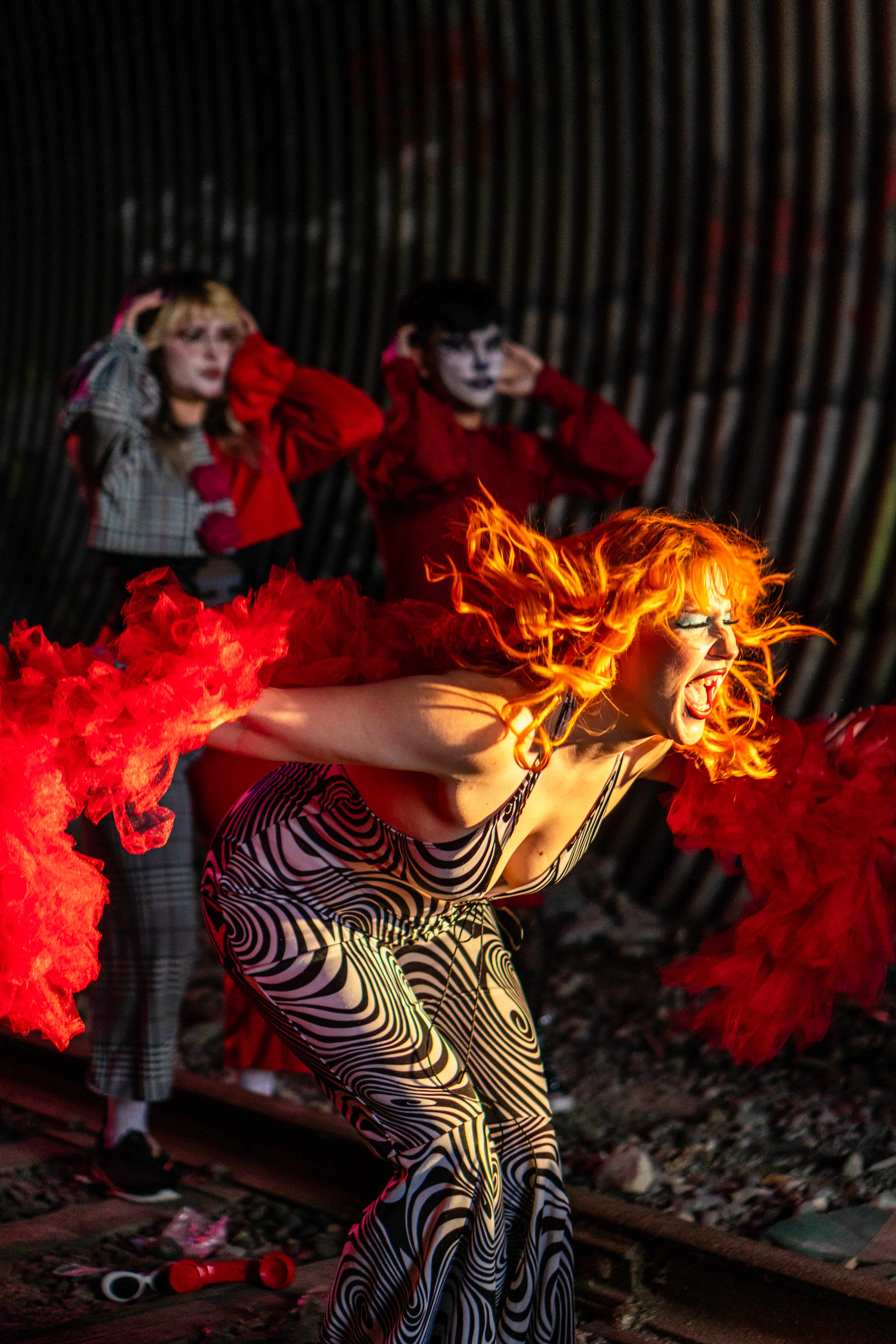 A performer with bright orange hair and dramatic makeup poses energetically in a black-and-white patterned outfit with flowing red fabric sleeves. Two blurred figures stand in the background under theatrical lighting, creating a bold, avant-garde performance scene with intense movement and emotion.