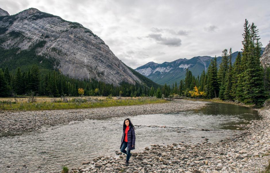 Carmen Aguirre walks along a river with mountains and trees in the background.