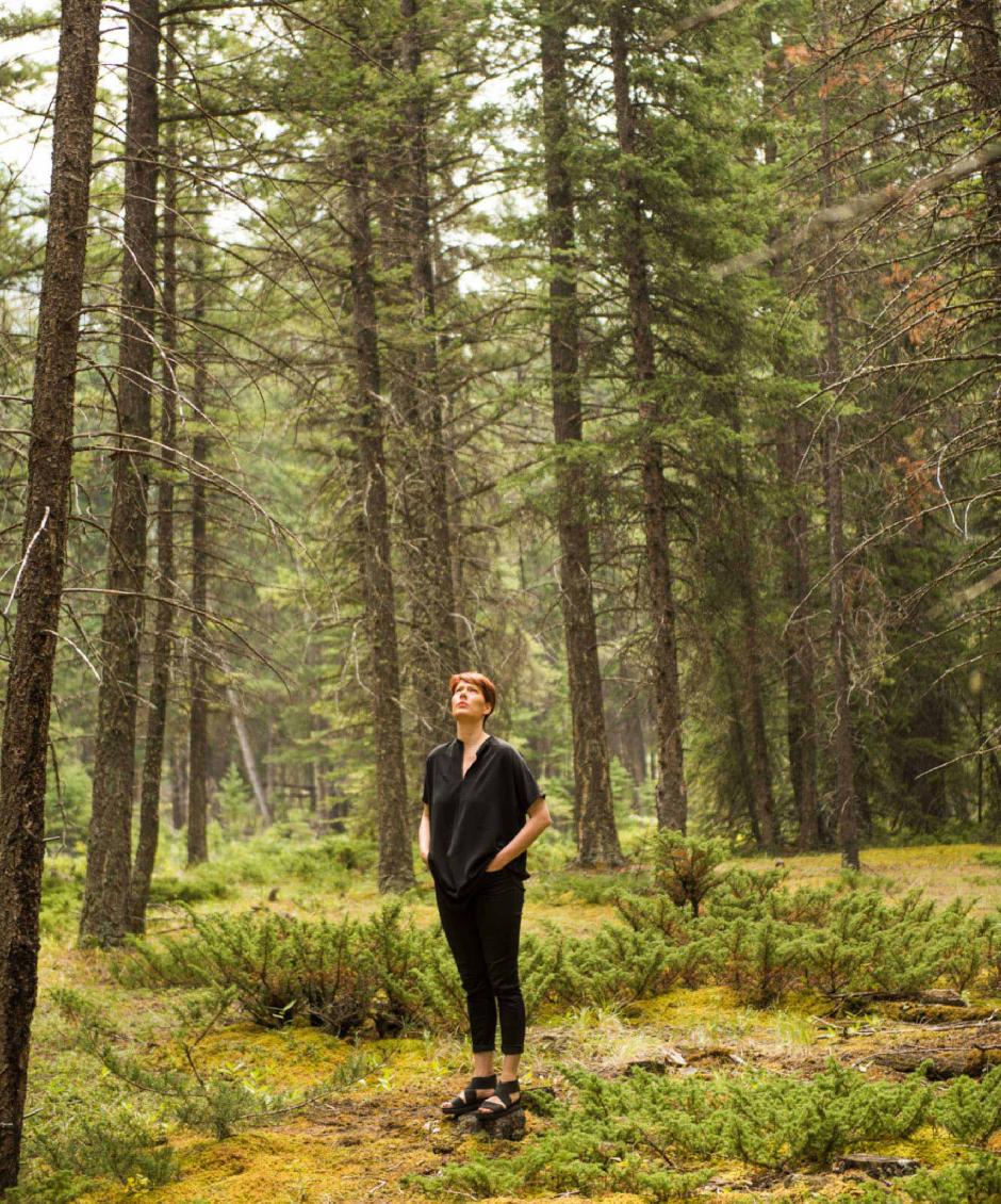 Emily Molnar stands in a forest surrounded by lush tall tress. 