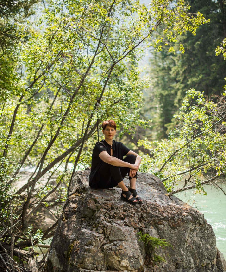 Emily Molnar posing on a rock sitting on the river front with green branches in the background.