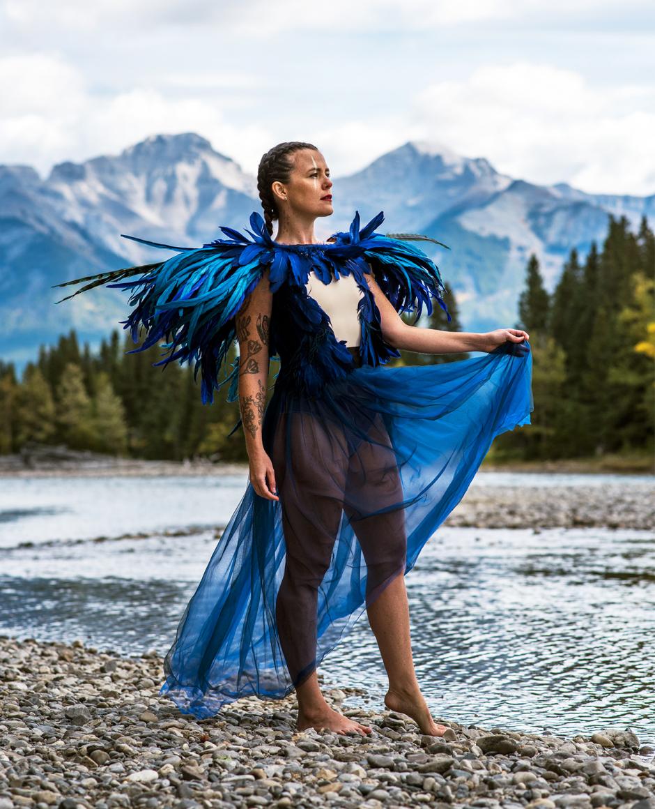 iskwē looks over a flowing river with mountain backdrop. 