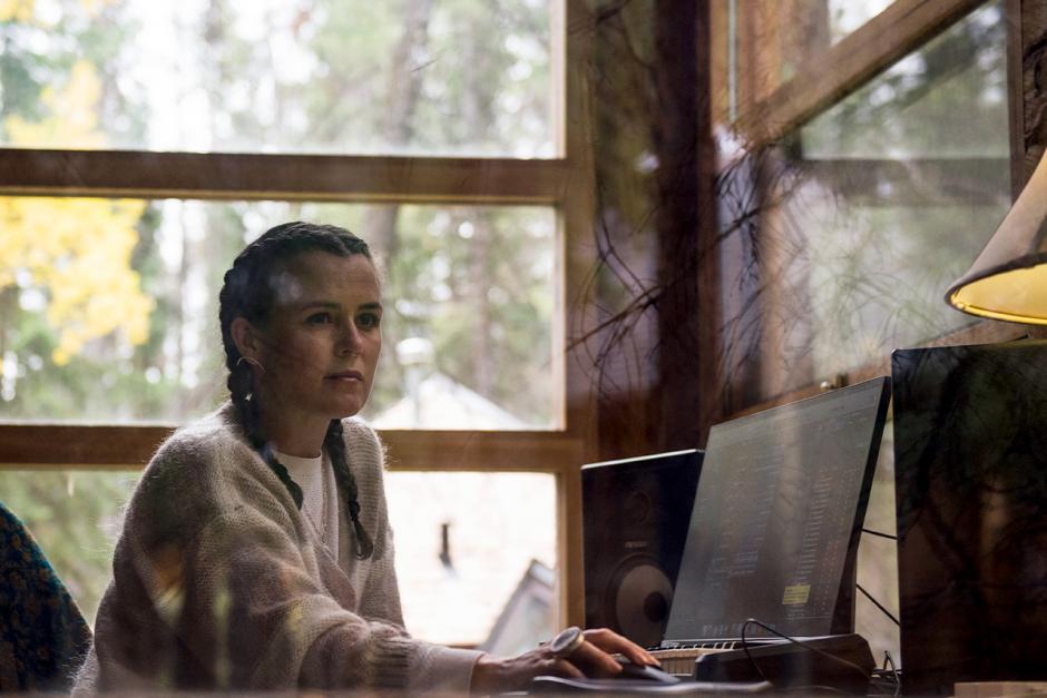 Musician iskwē works inside of of a studio with windows surrounding her.