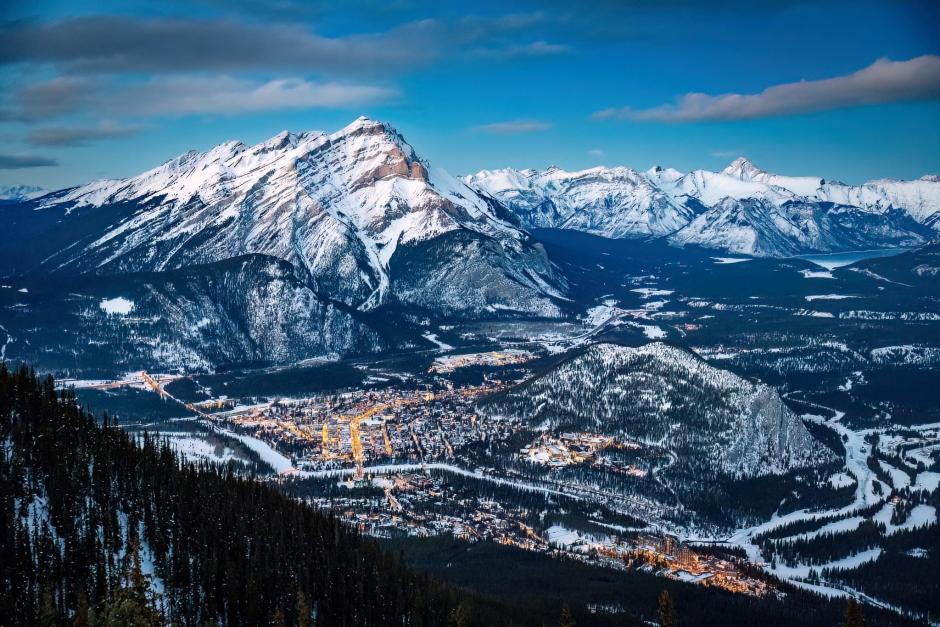 A snow covered Banff is seen from the Sulphur Mtn Gondola above the town to the South-West. It's a cool blue evening and in the background snow peaks line the horizon.