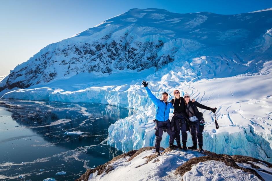 Three people stand upon a rock outcrop on the shore of Antarctica. Behind a small glacier capped peak rises above calm black waters. They look excited to be there on a sunny beautiful day. 