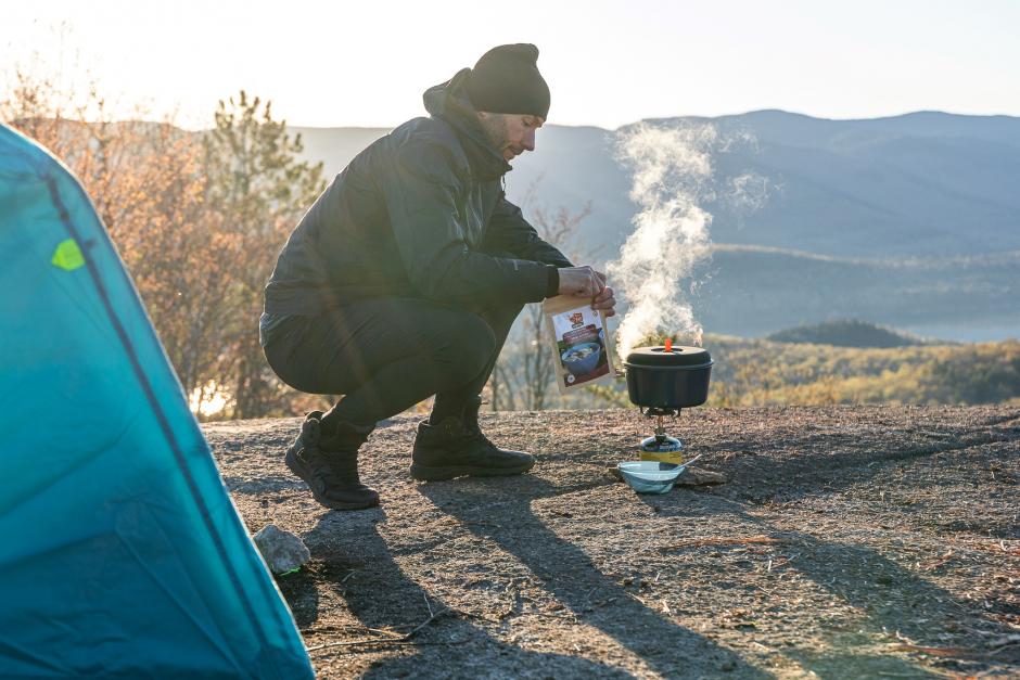 A man crouches by his tent holding a bag of Happy Yak dehydrated food and boiling water on a camp stove. His camping location has view over a large river or lake and small rolling mountains.