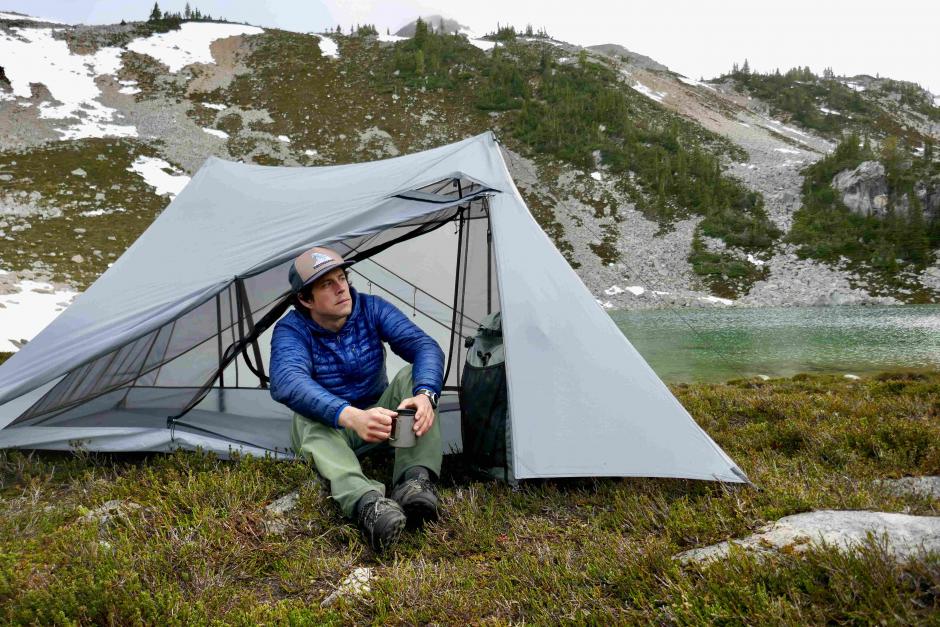 A man holding a coffee mug sits in a unique looking dyneema tent in the mountains. The tent is on a patch of grass by a small pond.