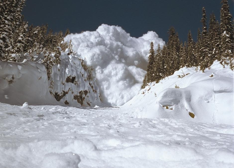 A very large avalanche is barreling down a narrow gully towards the camera, it is a blue sky above.
