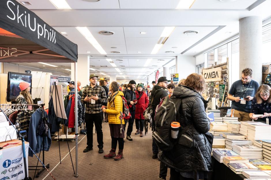 A large room is filled with numerous exhibitors interacting with the public. Books, clothing, and signage take up space on the visible tables.