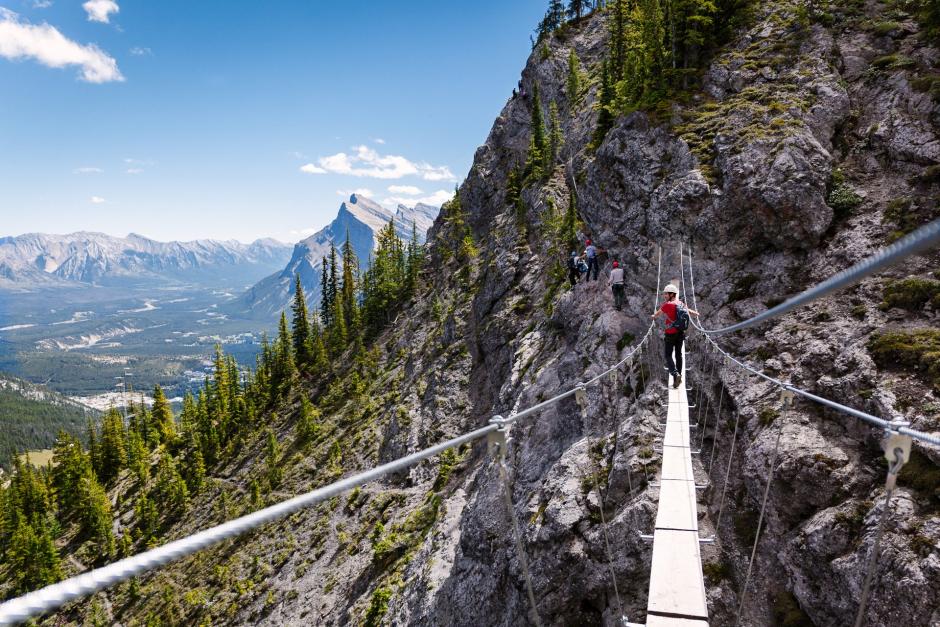 A man walks a via ferrata bridge between mountain peaks at Mt. Norquay above the town of Banff,