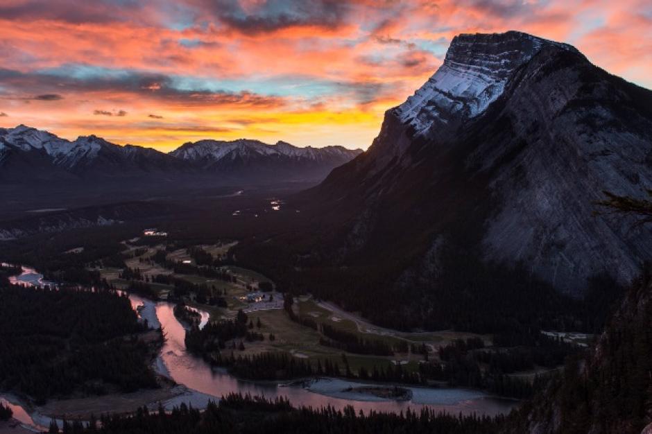 Sun rises behind Mt. Rundle as seen from Tunnel Mountain, the sky has fiery orange and bright pink clouds.
