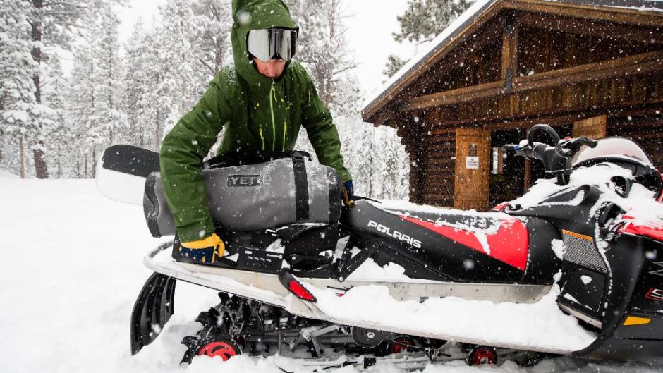 A man loads a Yeti duffle onto the back of a Snowmobile in front of a wood cabin.