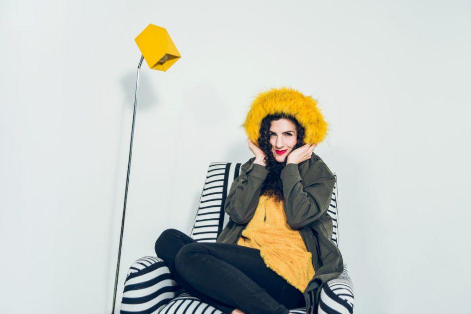 Laila Biali is wearing a bright yellow shirt and matching fur hat, while sitting on a black and white striped armchair, next to a lamp with a yellow lampshade.