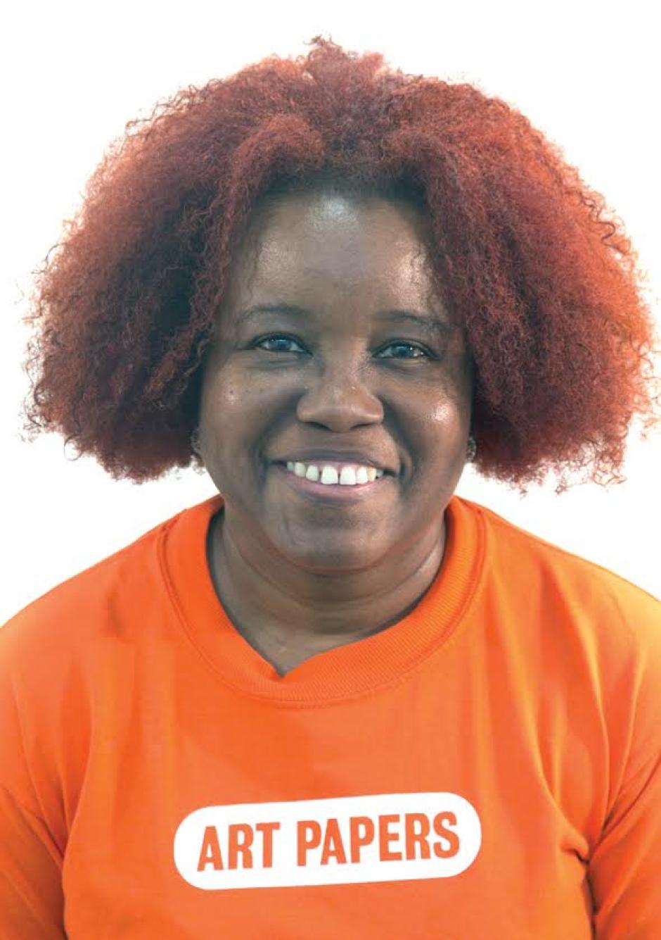 Fatimah Tuggar Looks straight forward wearing an orange shirt that reads "Art Papers"