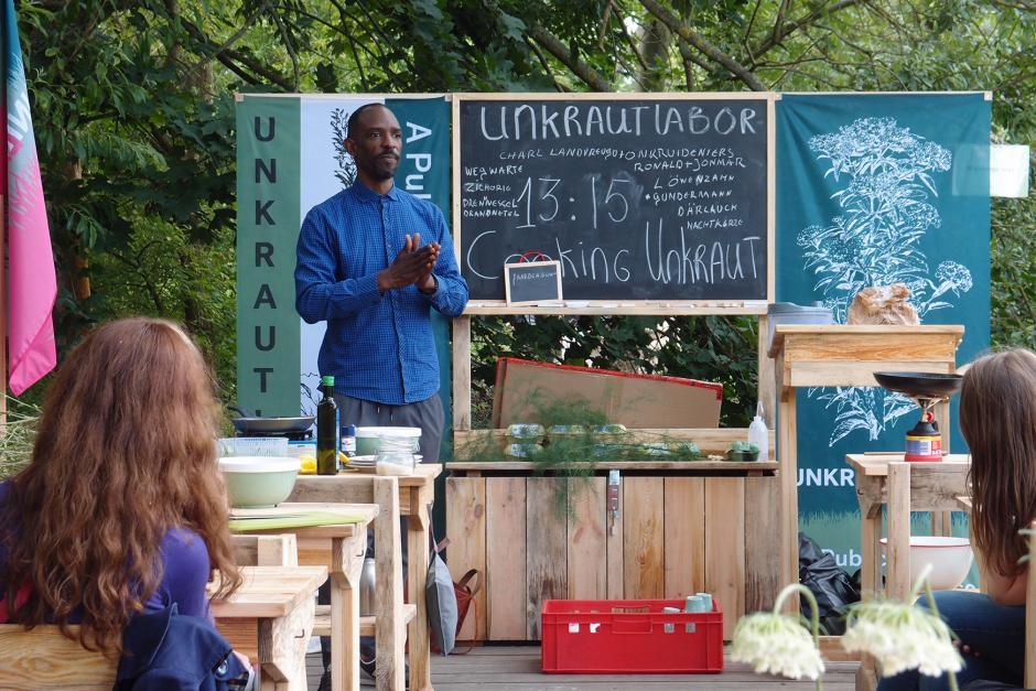 A man stands at the front of an informal outdoor classroom lecturing on Unkraut