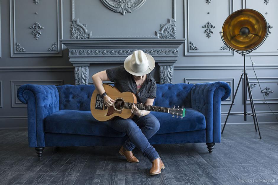 Irish sits on a blue felt couch wearing a broad rimmed hat playing her guitar.