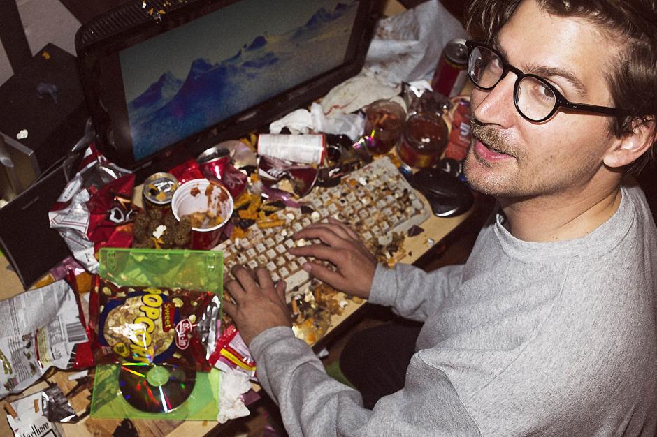 Visual Artist Jon Rafman sits with his hands on a filthy desktop and keyboard covered in fast-food items.