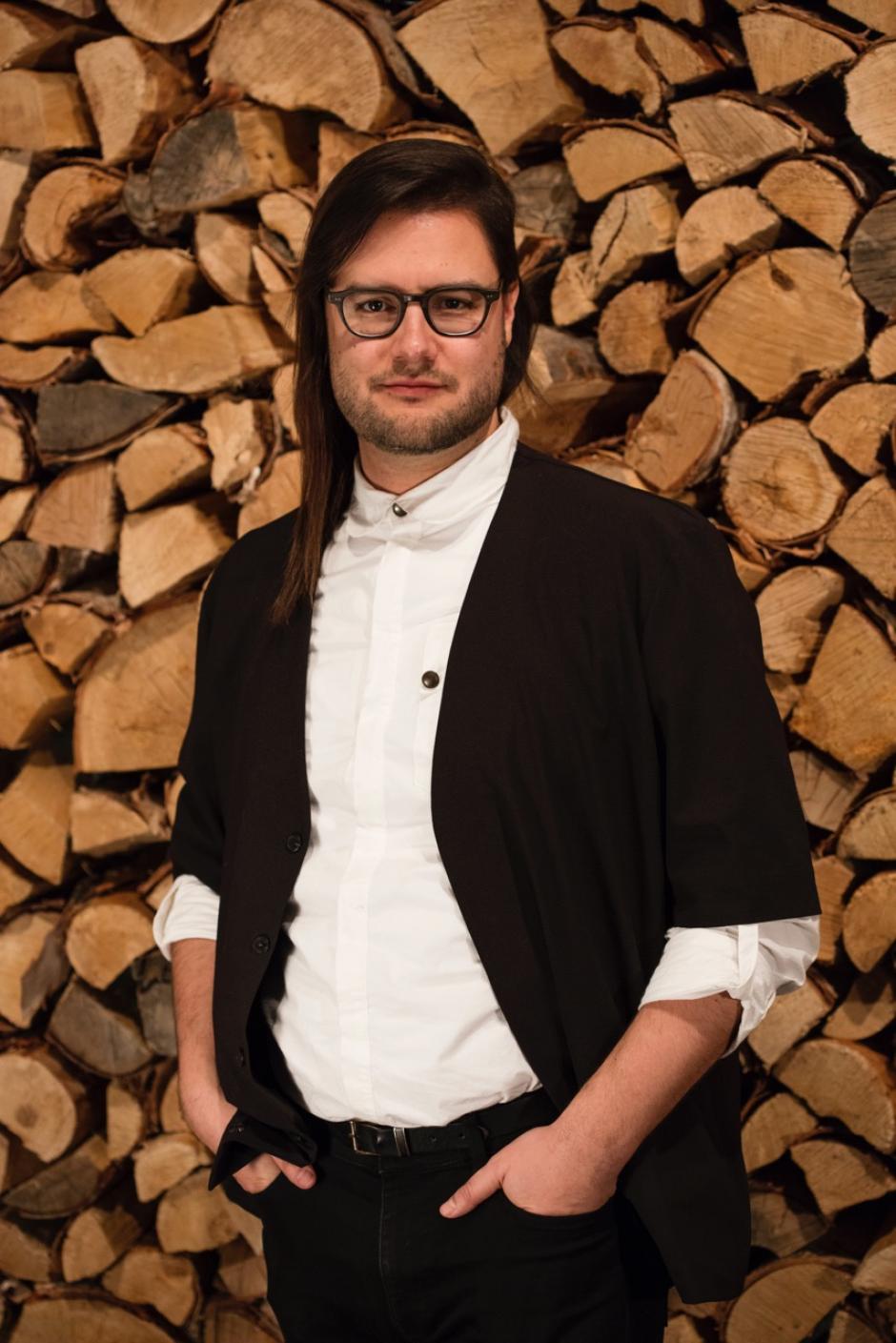 Visual Artist John G. Hampton stands in front of a wall of split logs.