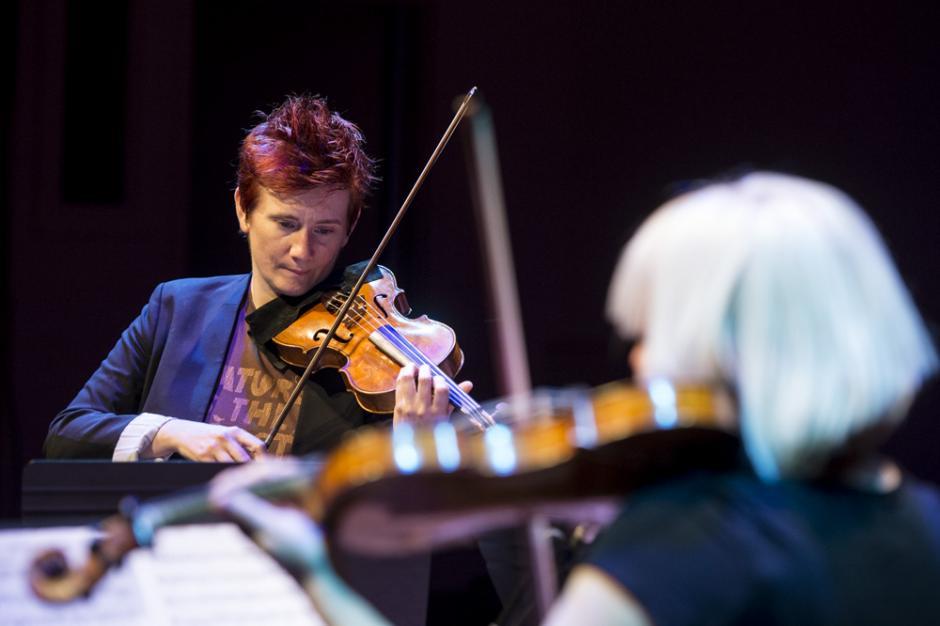 Two women face each other playing violin, the one head-on from the photographer is in focus.